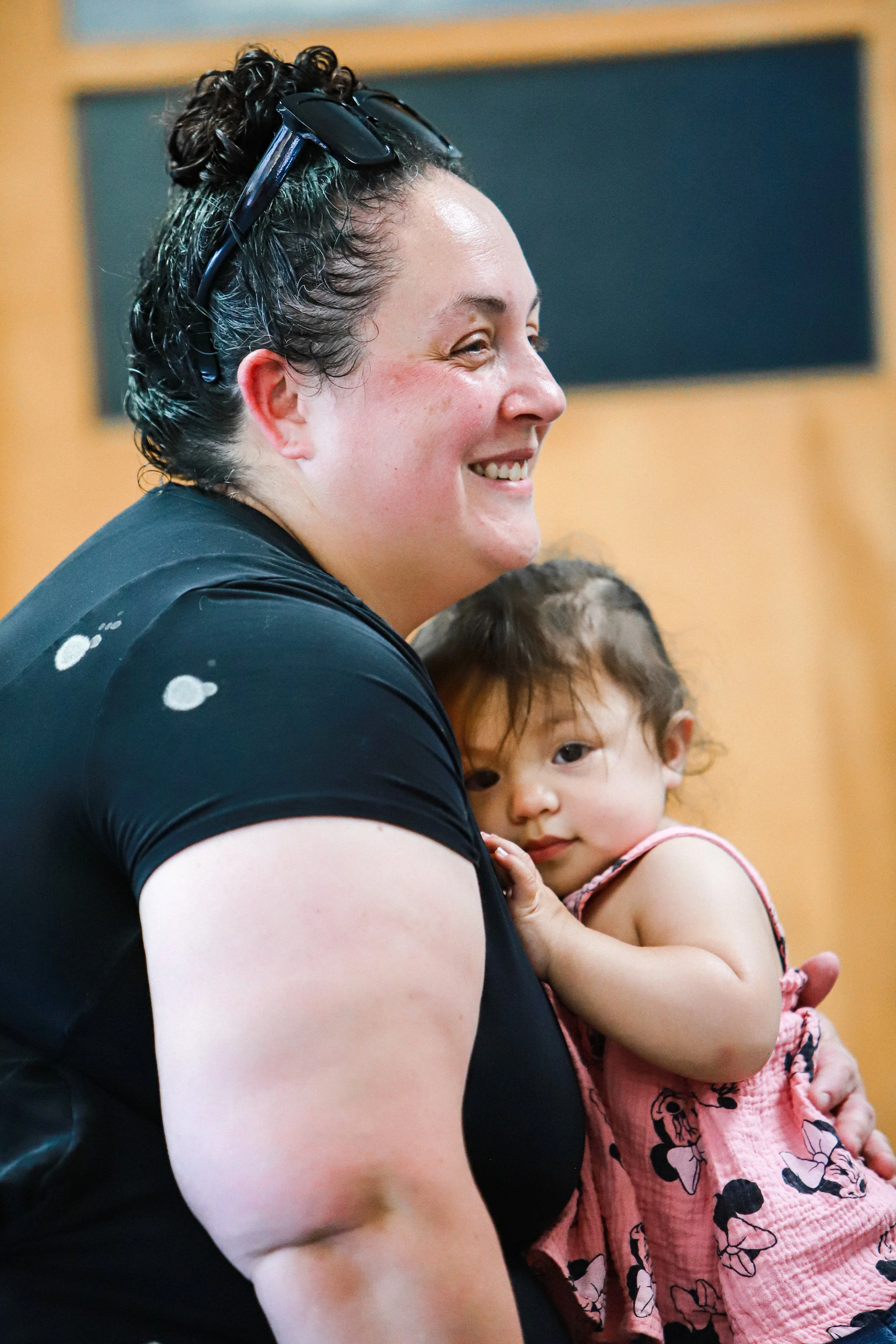 A woman with glasses on her head smiling while holding a young girl in her arms, inside a room with a wooden wall in the background.