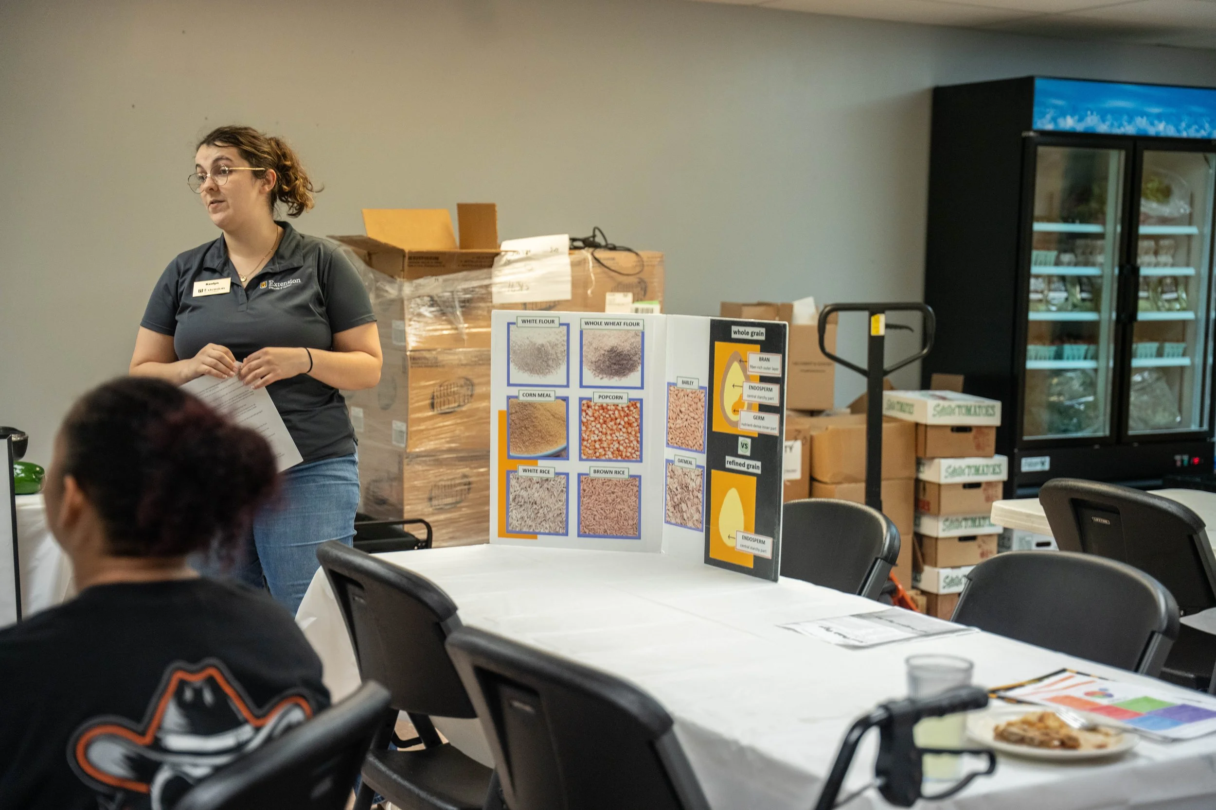 A woman giving a presentation in a room with a table, chairs, and informational display boards showing grains and food products.