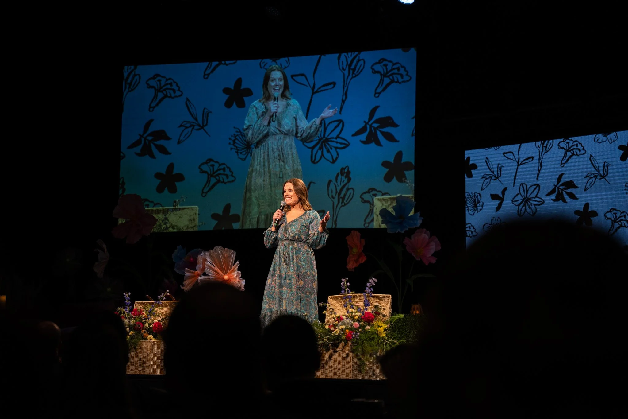 A woman in a patterned dress speaks into a microphone on stage, with a large screen behind her displaying her image and a background of black-outlined flowers and leaves. The stage is decorated with flowers and woven baskets.