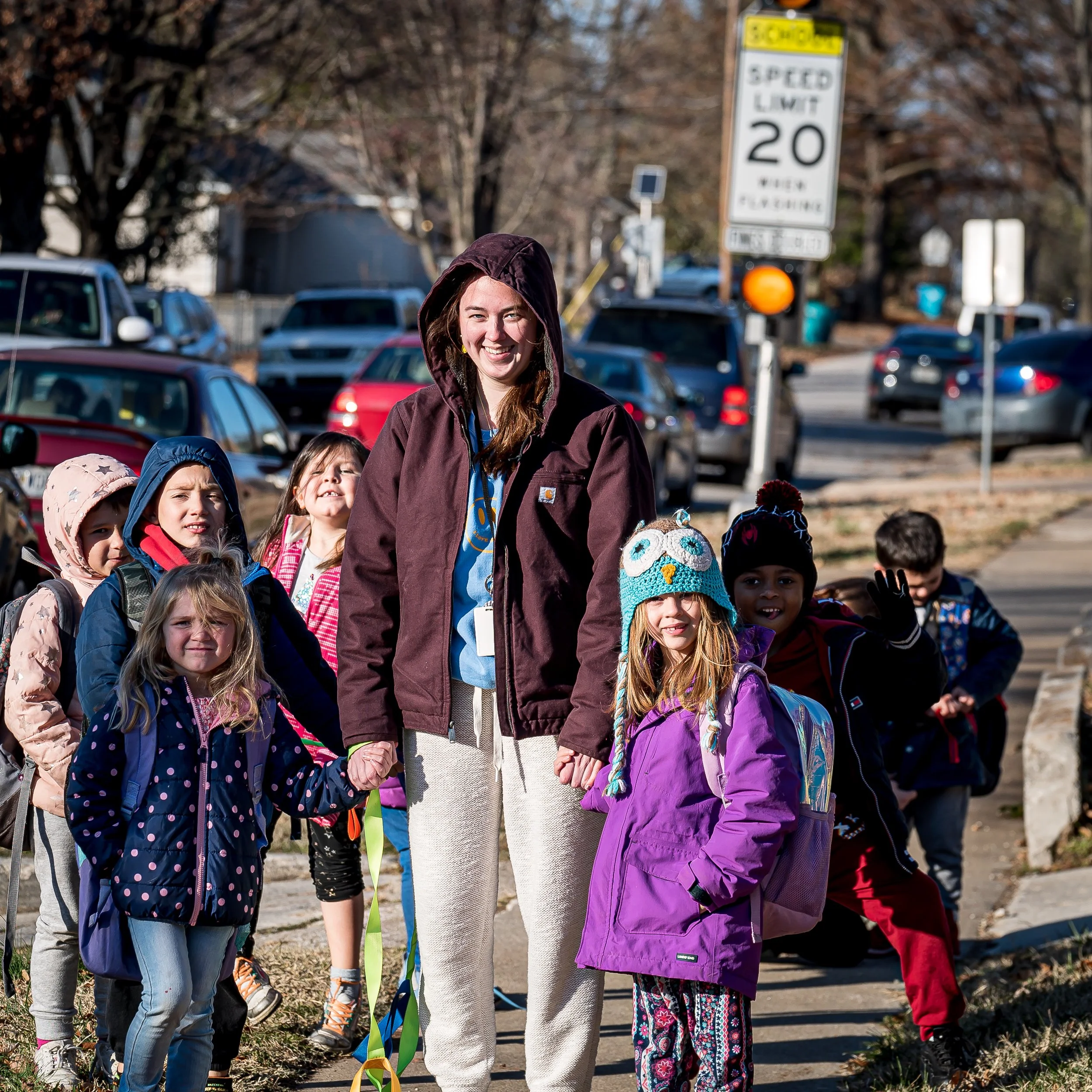 A woman leading a group of children on a sidewalk, holding a ribbon, with parked cars and a street sign in the background.