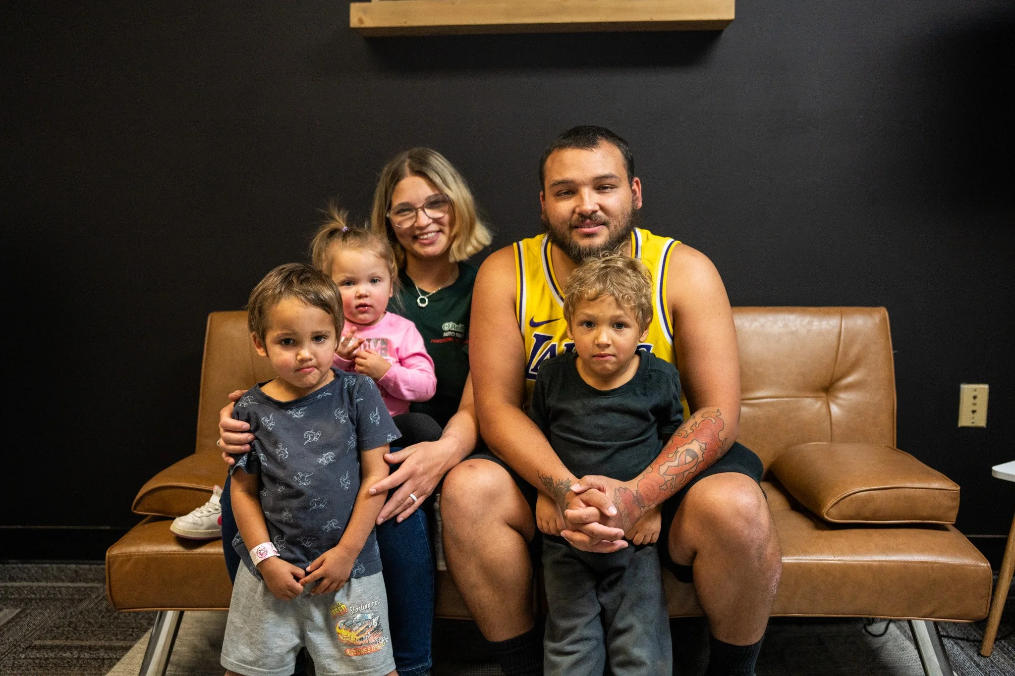 A group of five people sitting on a brown leather couch against a black wall. The group includes a woman with glasses, a man in a yellow Lakers jersey, two boys, and a girl. The woman is smiling, and the children are looking at the camera with neutral expressions.