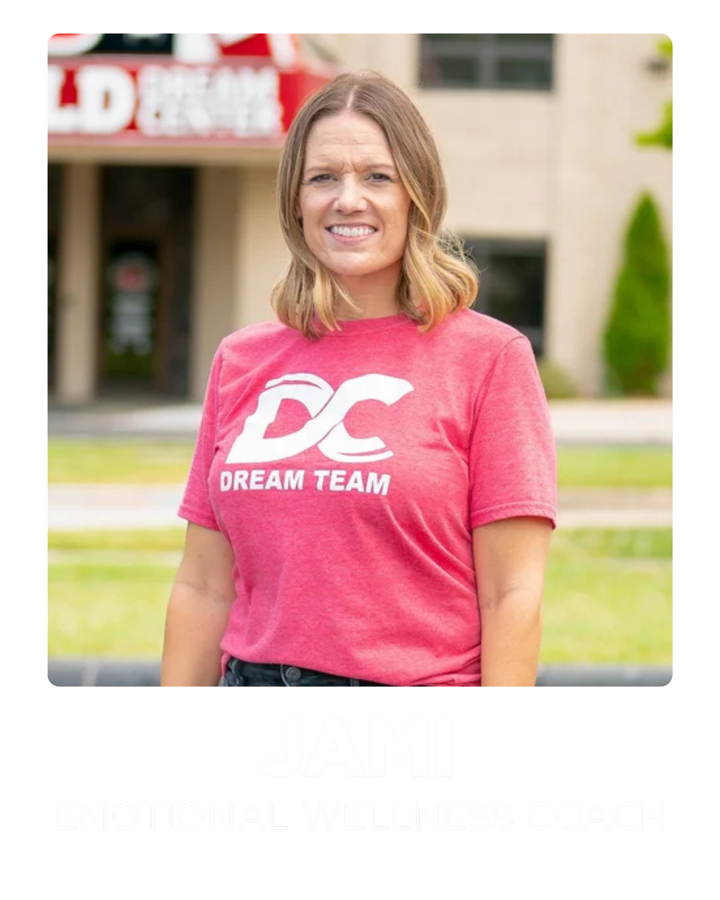 A woman with shoulder-length blonde hair wearing a pink t-shirt with white text and logo, standing outdoors in front of a blurred background of a building and greenery.