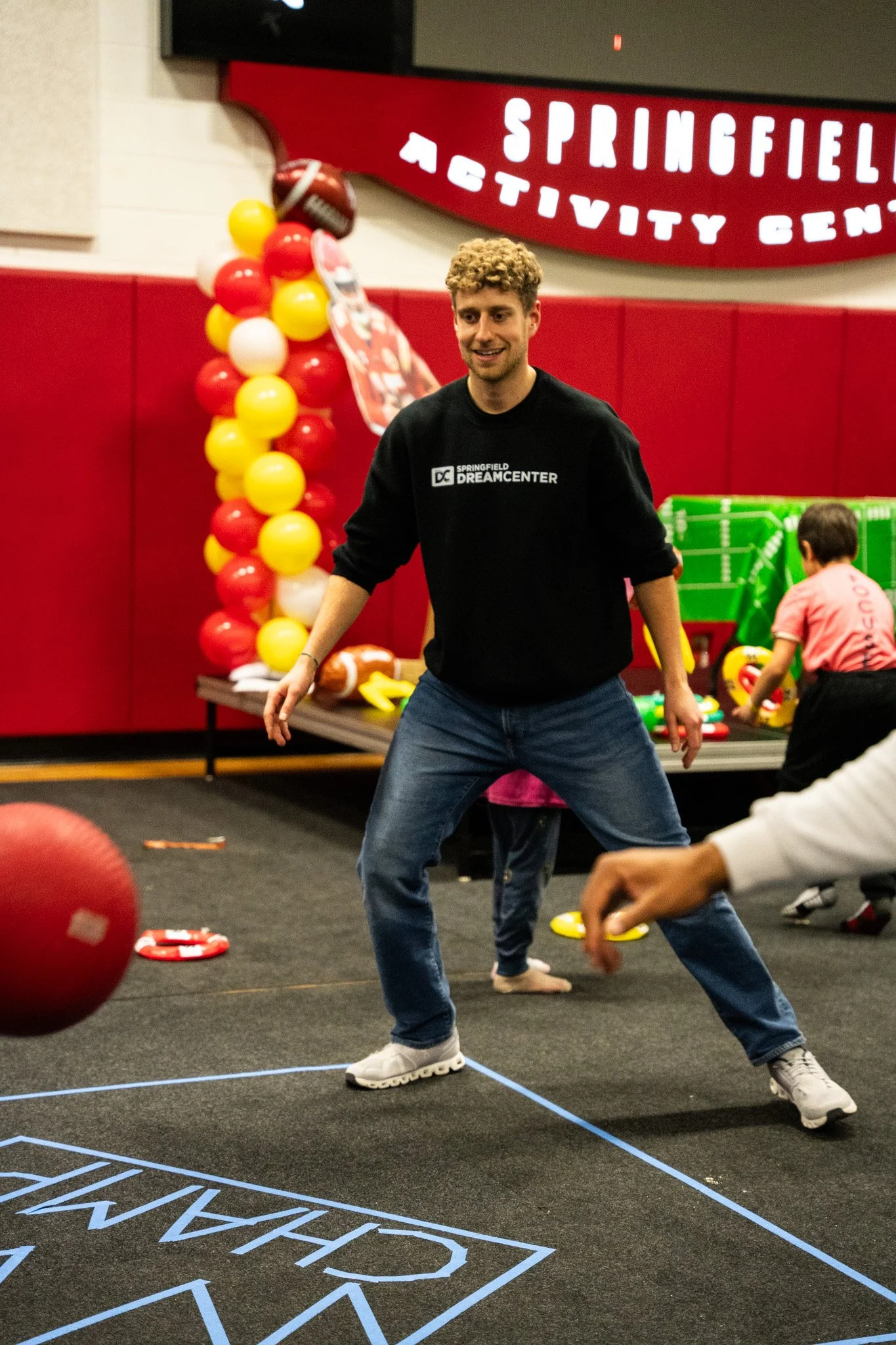 A young man with curly hair in a black sweatshirt and jeans plays a game at Springfield DreamCenter, with balloons and children in the background.