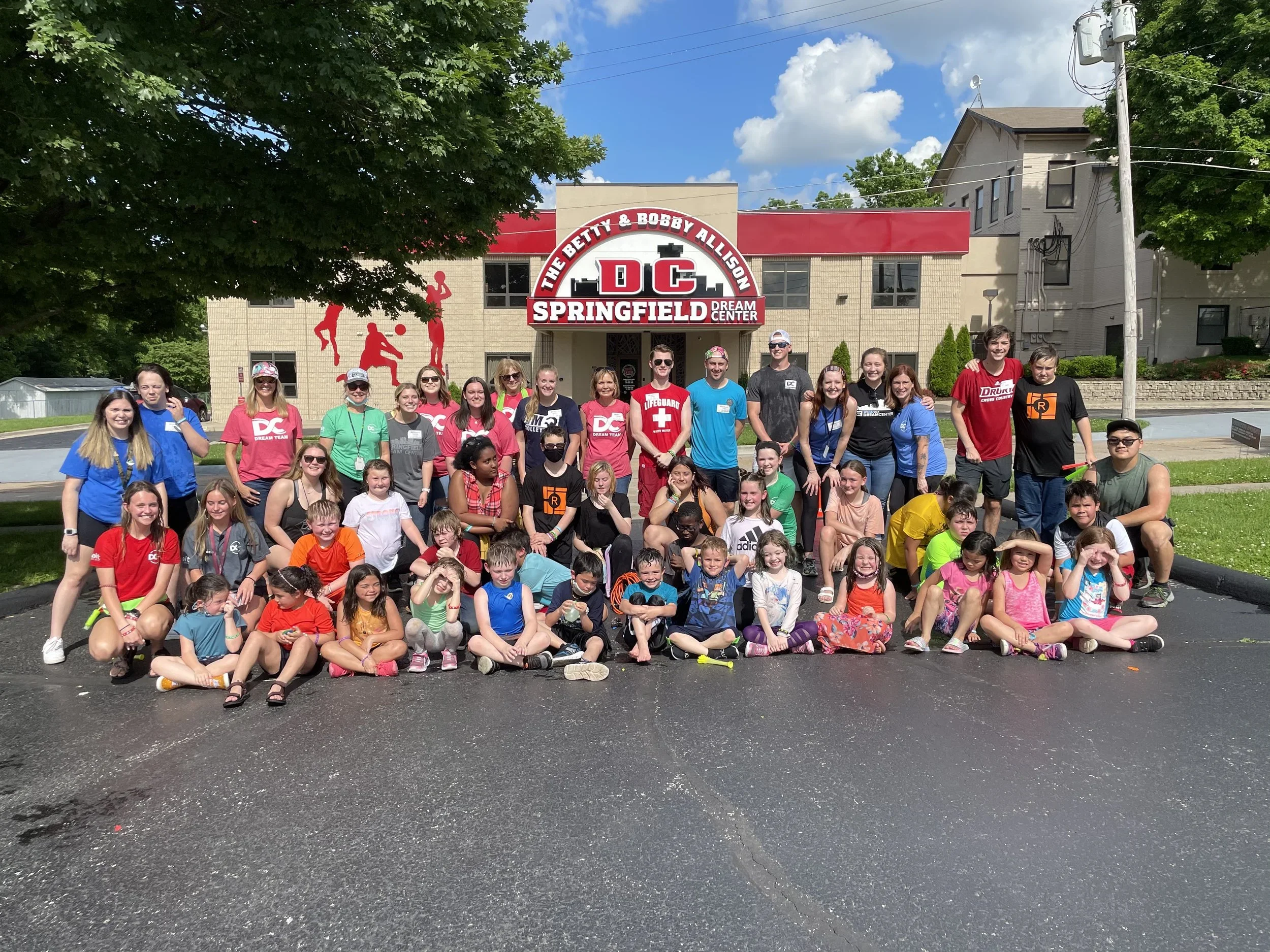 A large group of children and adults posing for a photo in front of the Springfield Dream Center building, with a sign that says 'The Betty & Bobby Allison DC Springfield Dream Center.' The group includes people of various ages, wearing colorful casual clothing, some with sunglasses and masks, with a clear sky and trees in the background.