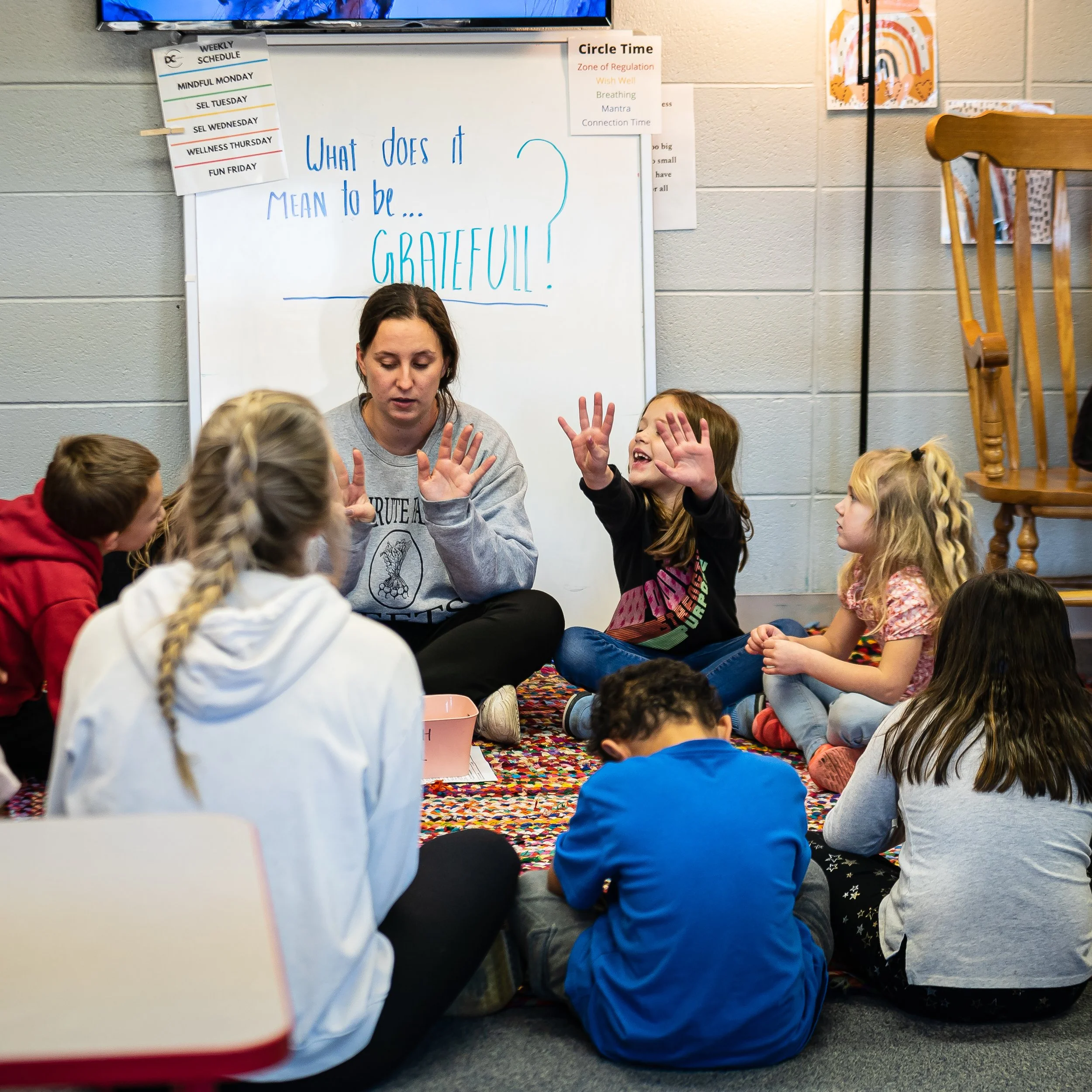 A group of children and a woman sitting on a colorful rug in a classroom, participating in a discussion about gratitude. The woman is speaking with her hands raised, and a girl next to her is raising her hands enthusiastically. A whiteboard behind them has the question, 'What does it mean to be... Grateful?' written on it.