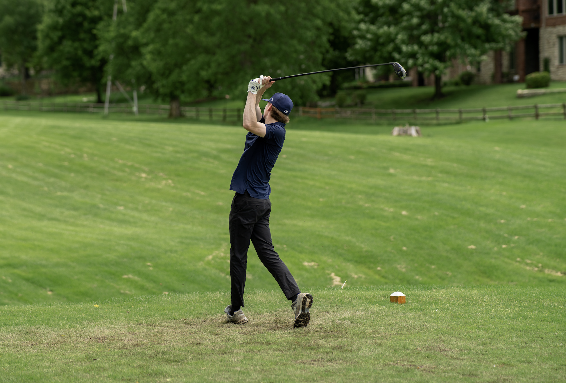 A man playing golf on a grassy course, swinging a golf club with a background of trees and houses.