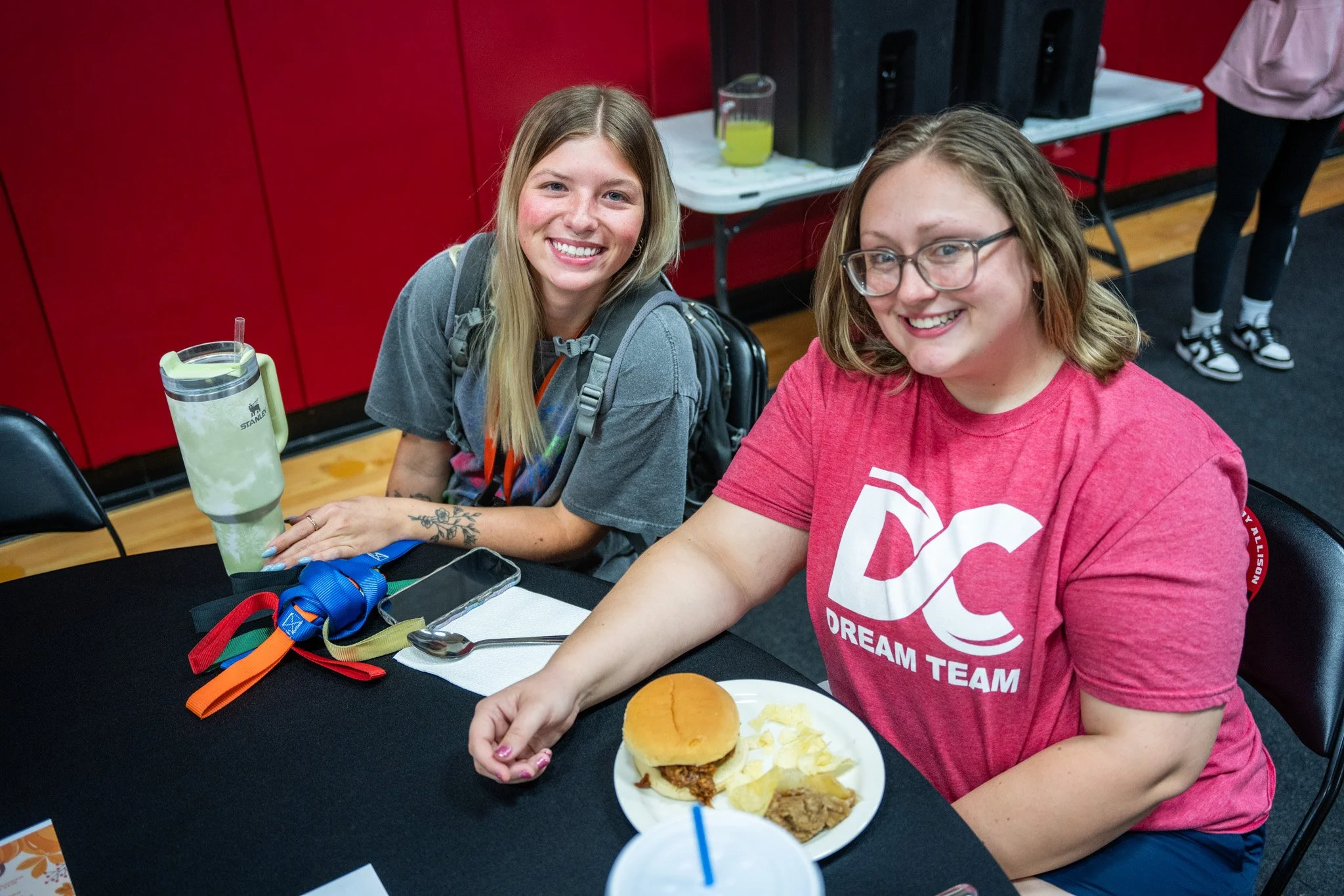 Two smiling young women sitting at a black table in a room with red walls, one with a tray of food including a burger and chips, and the other with a large water bottle, various personal items, and a phone.