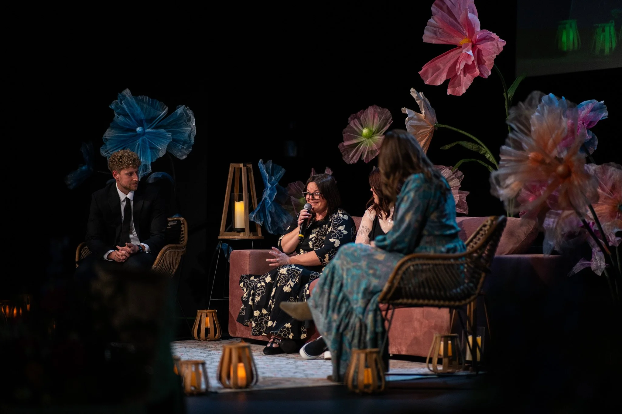 Four people sitting on stage with floral decorations, including large pink and blue flowers, during a panel or discussion event.