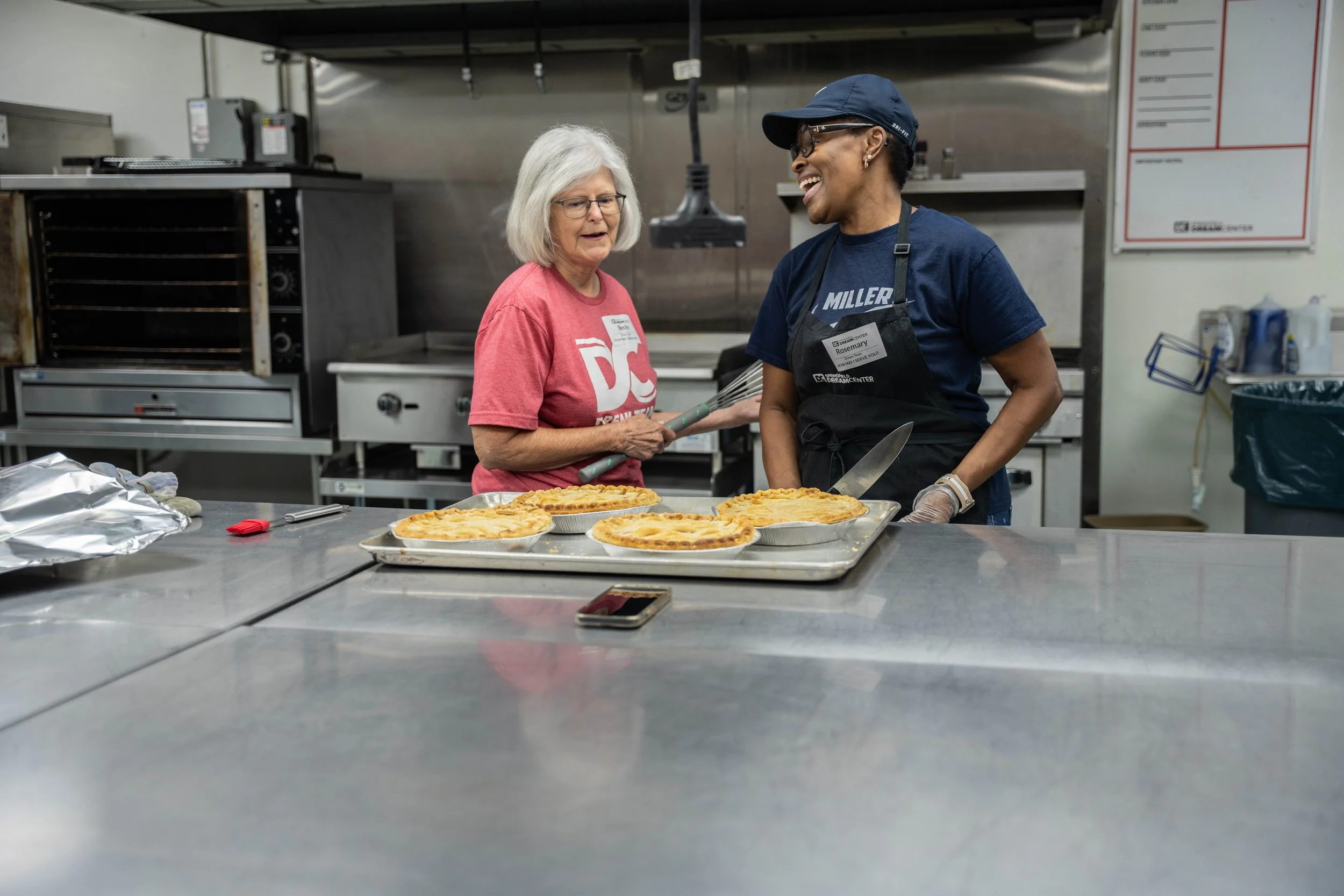 Two women, one elderly and one middle-aged, laughing and talking in a commercial kitchen, with four cooked pies on a tray in front of them.