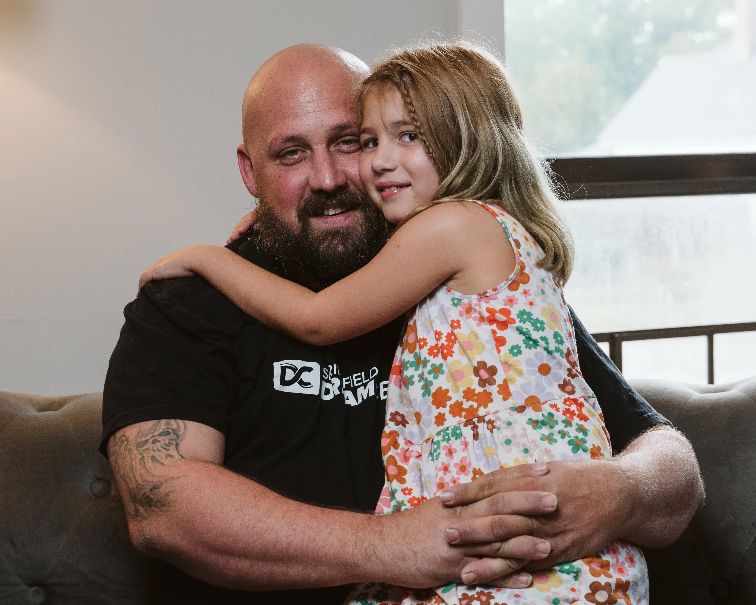 A man with a beard and a tattoo on his arm is hugging a young girl with long, light brown hair and braids. The girl is wearing a sleeveless floral dress and is embracing the man. They are sitting on a gray couch near a window.