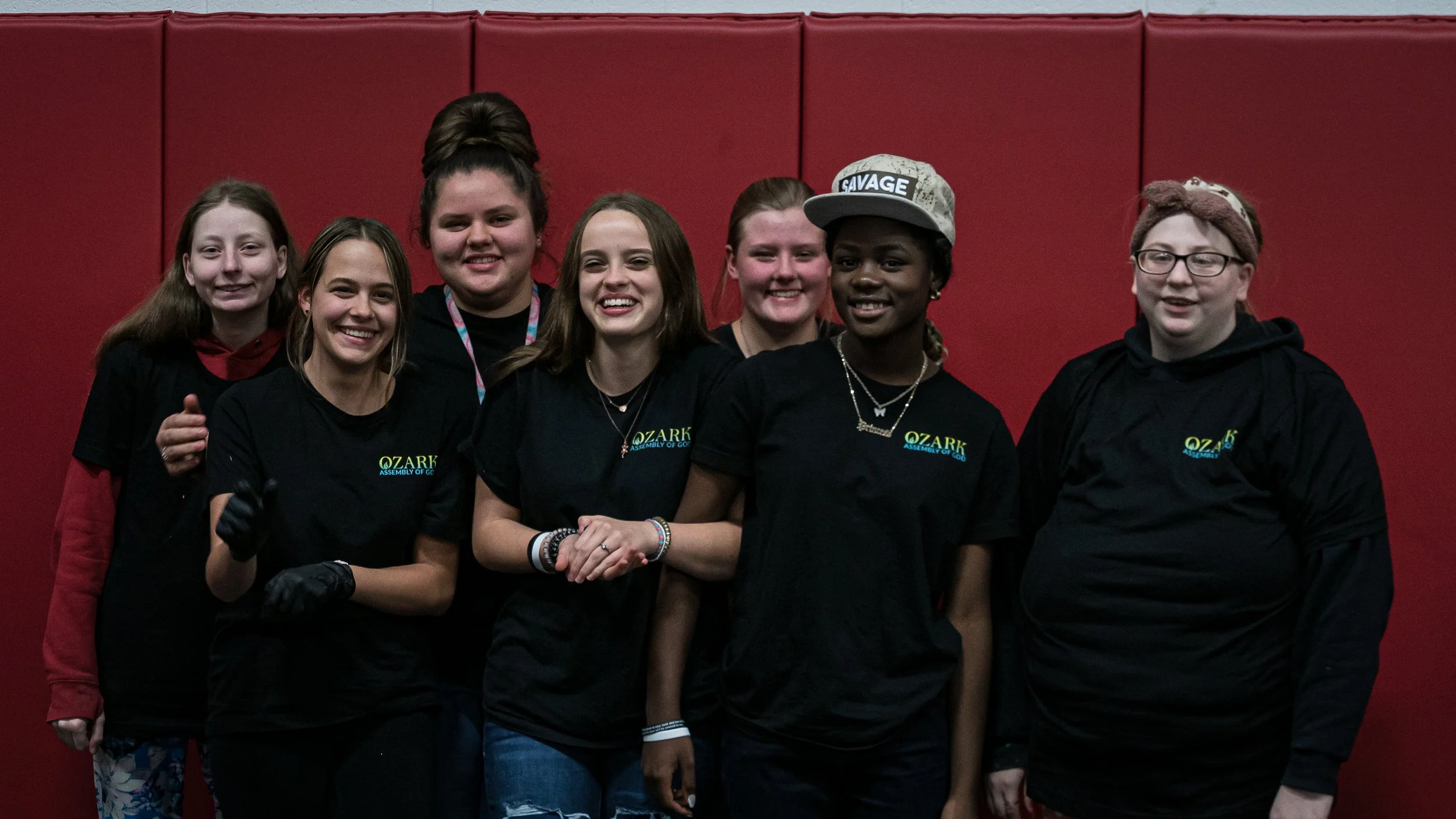Group of seven young girls wearing black 'Ozark Assembly of God' T-shirts, standing against a red background, smiling for the camera.