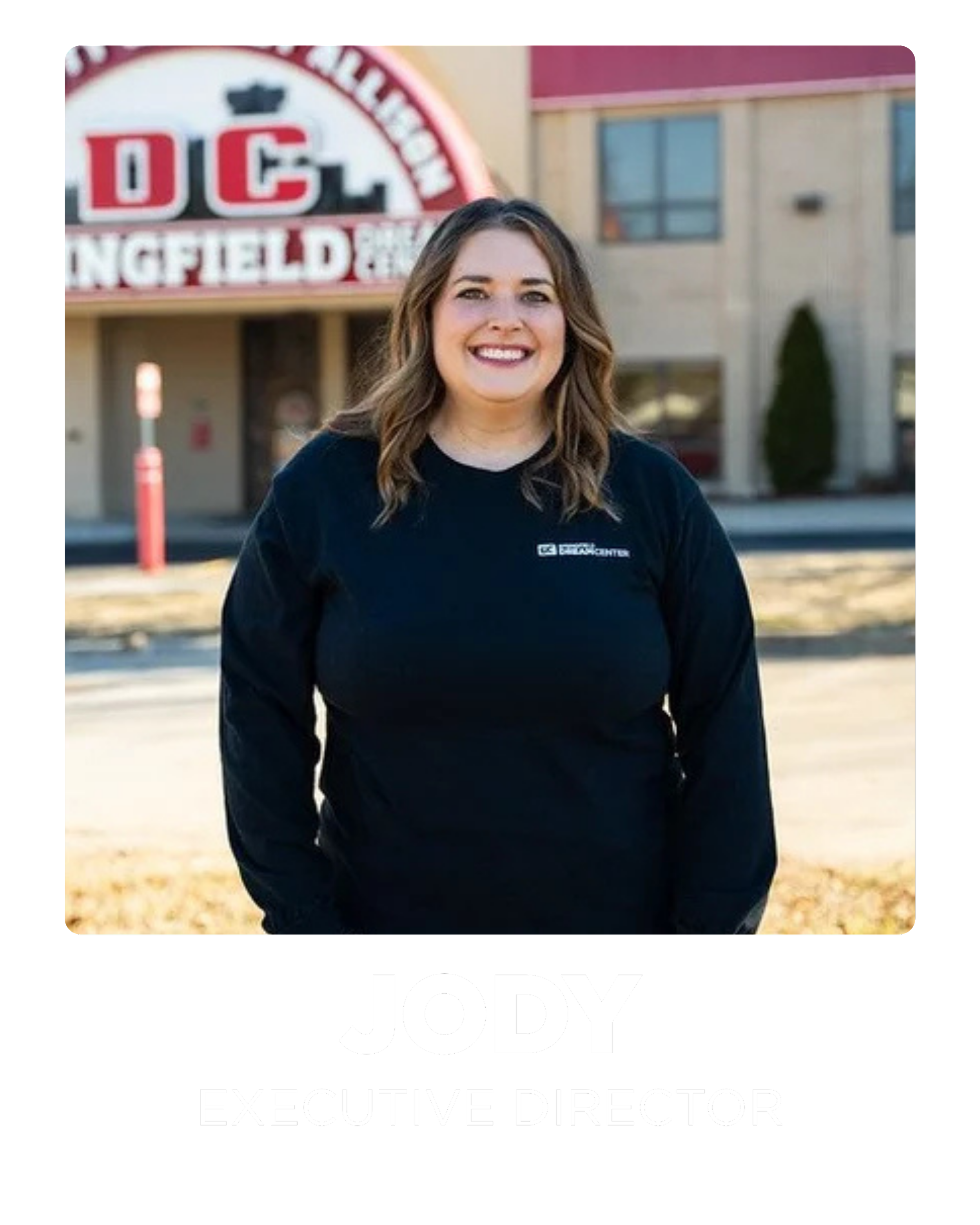 A woman with shoulder-length brown hair smiling, standing outdoors in front of a building with a sign that reads 'DCL' and 'Springfield'. She is wearing a black long-sleeve shirt with a logo and text. The bottom of the image has a black banner with her name 'Jody' and the title 'Executive Director' in white text.