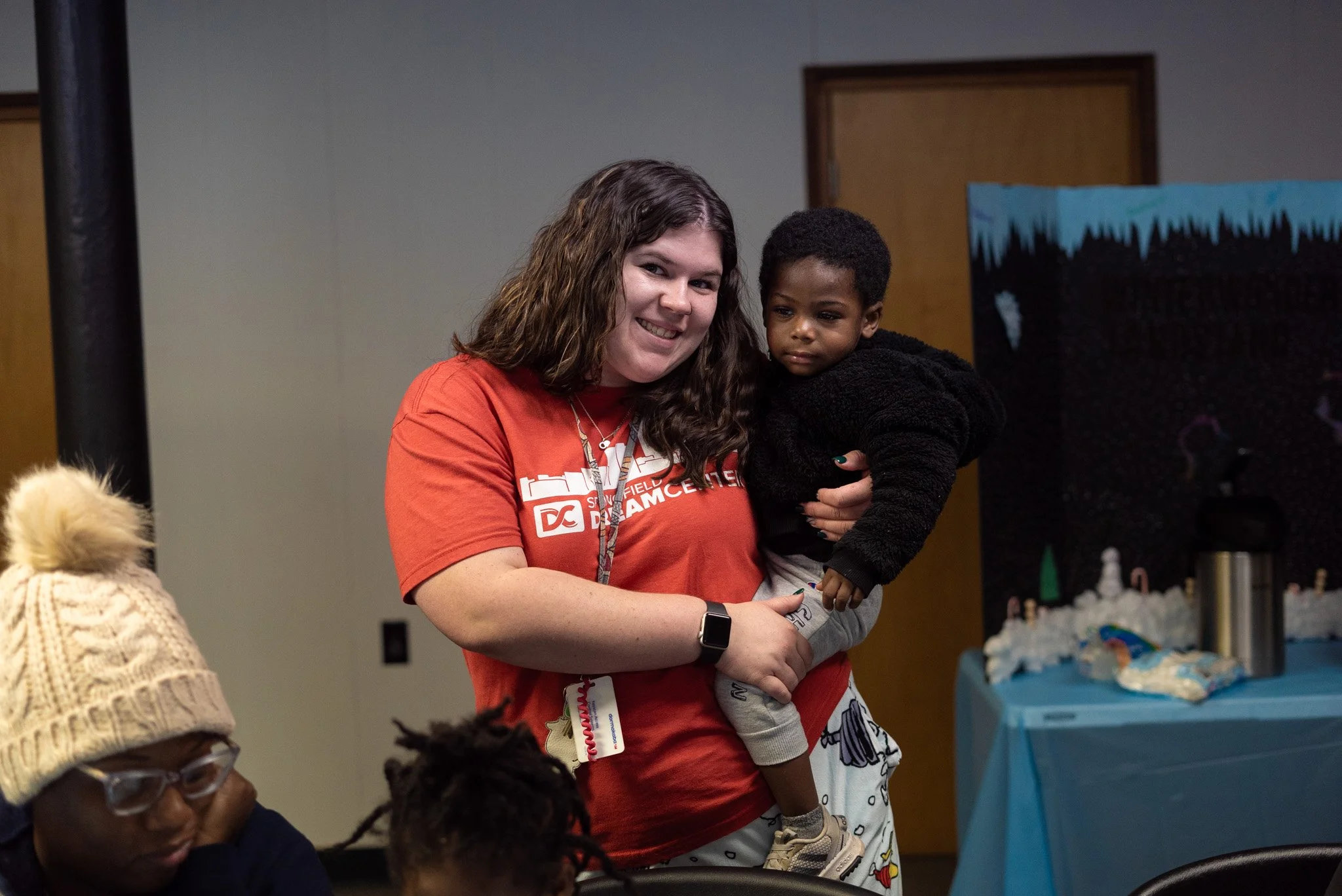 A woman with curly brown hair smiling while holding a young boy, both standing indoors near a table with a blue tablecloth and a winter-themed display. The woman is wearing a red t-shirt and a smartwatch, and the boy is wearing a black fuzzy jacket a