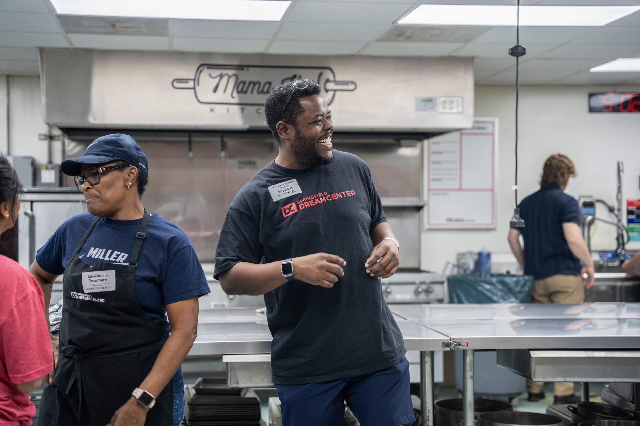 People working in a kitchen, wearing name tags, with one person smiling and engaging in conversation.