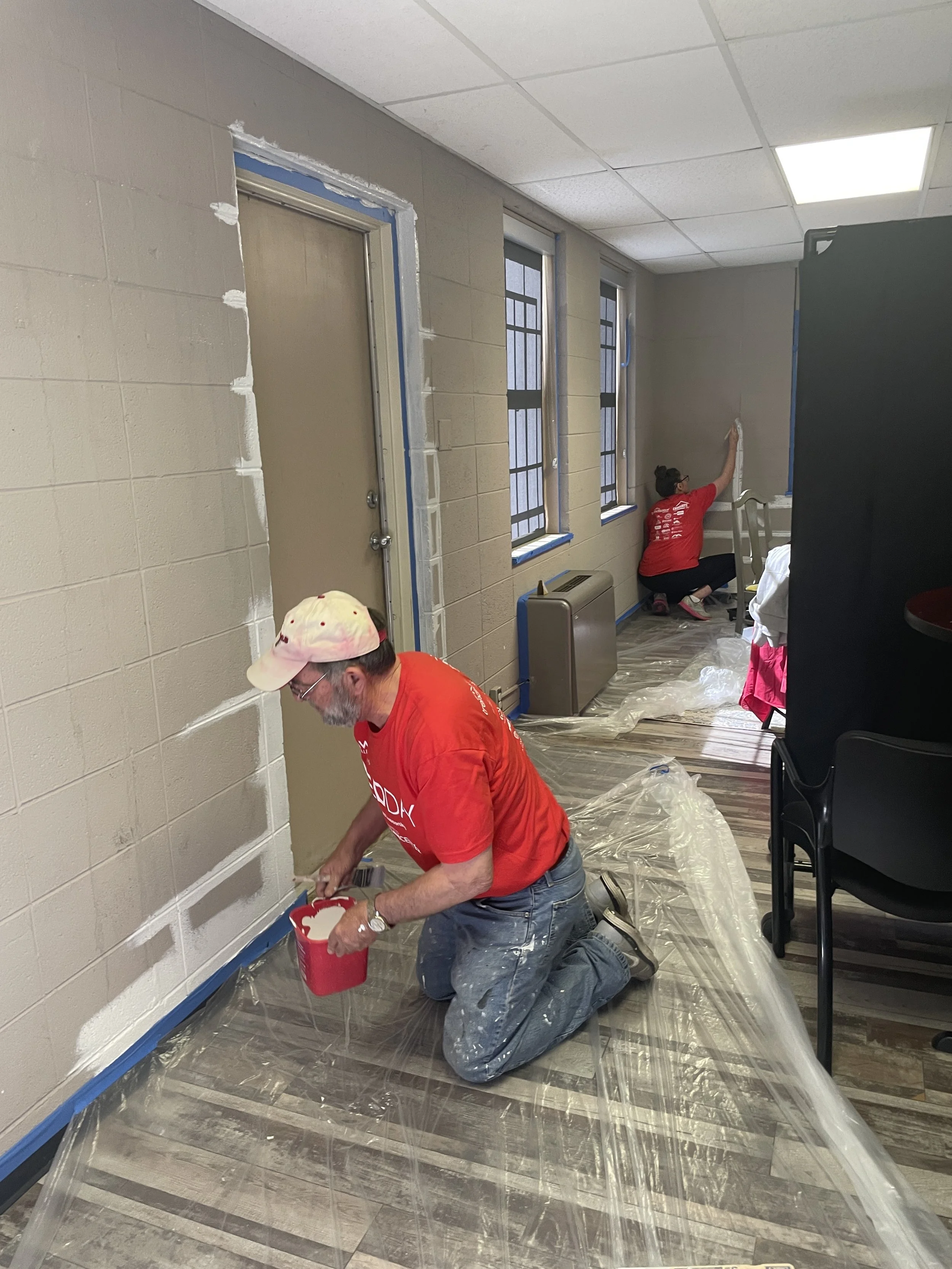 Two people painting a wall in a room with plastic covering the floor, windows, and door frame, while kneeling on the floor and using brushes.