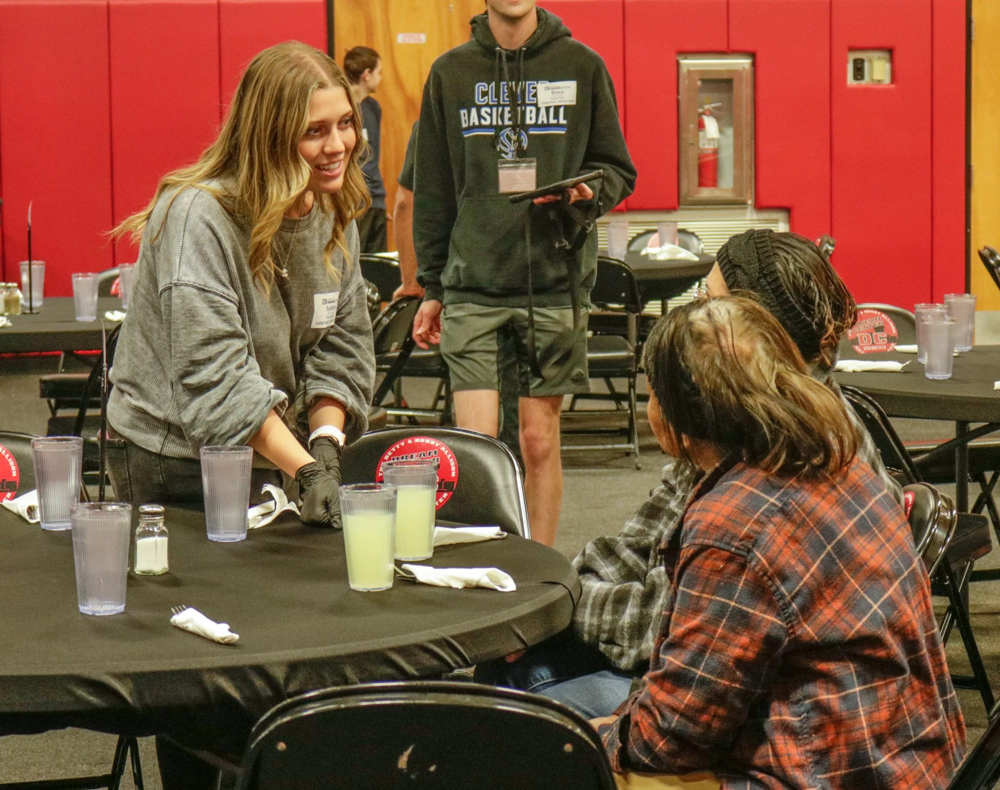 A woman with blonde hair, wearing a gray oversized sweater, smiling and talking to a person sitting at a table in a restaurant or event setting. The table has glasses, utensils, and condiments. A man in a green hoodie with a Cleveland basketball logo