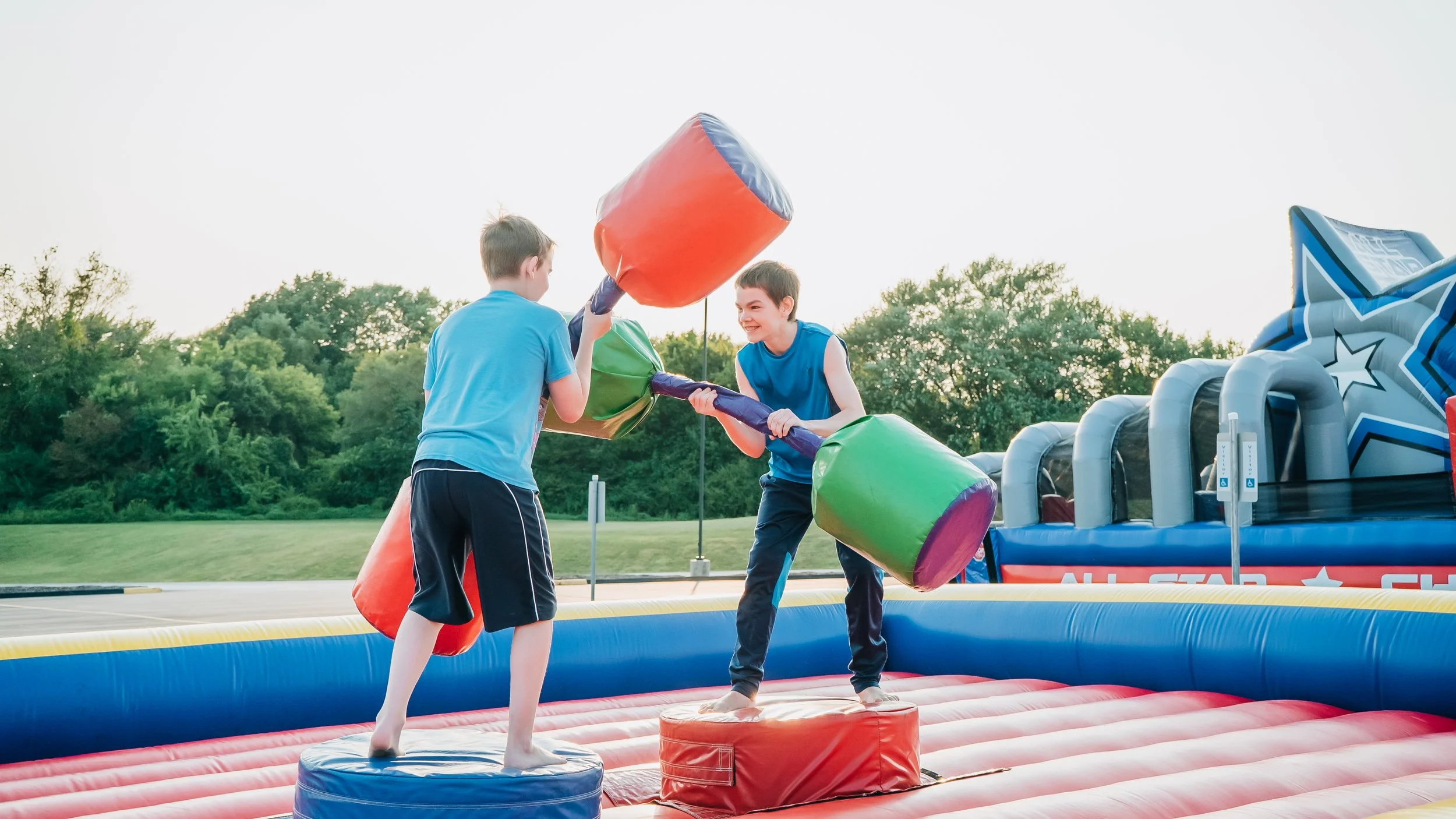 Two boys playing on an inflatable jousting game at a park or fair, with a grassy field and trees in the background.