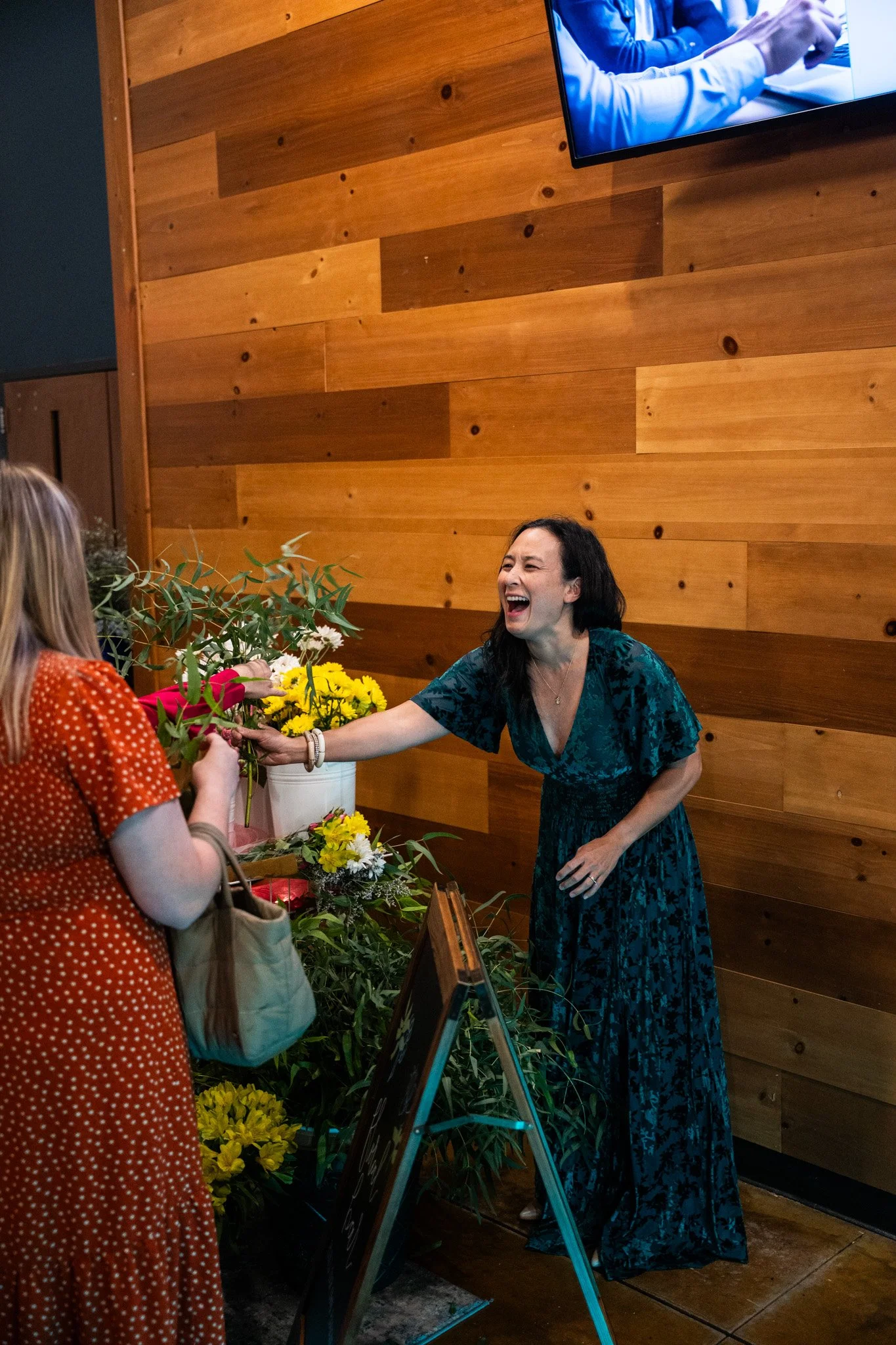 A woman in a blue dress smiling and laughing while exchanging flowers with another woman in an orange dress at an indoor event with a wooden wall background.