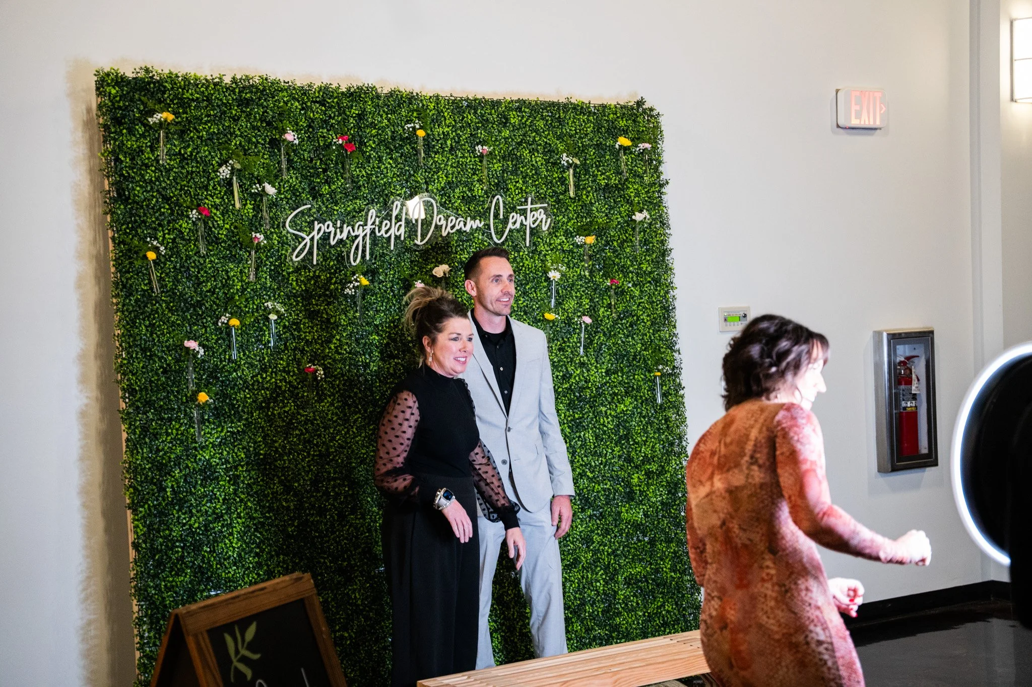 A man and a woman posing for a photo at the Springfield Dream Center event, standing in front of a green wall decorated with flowers, with another woman in the foreground.