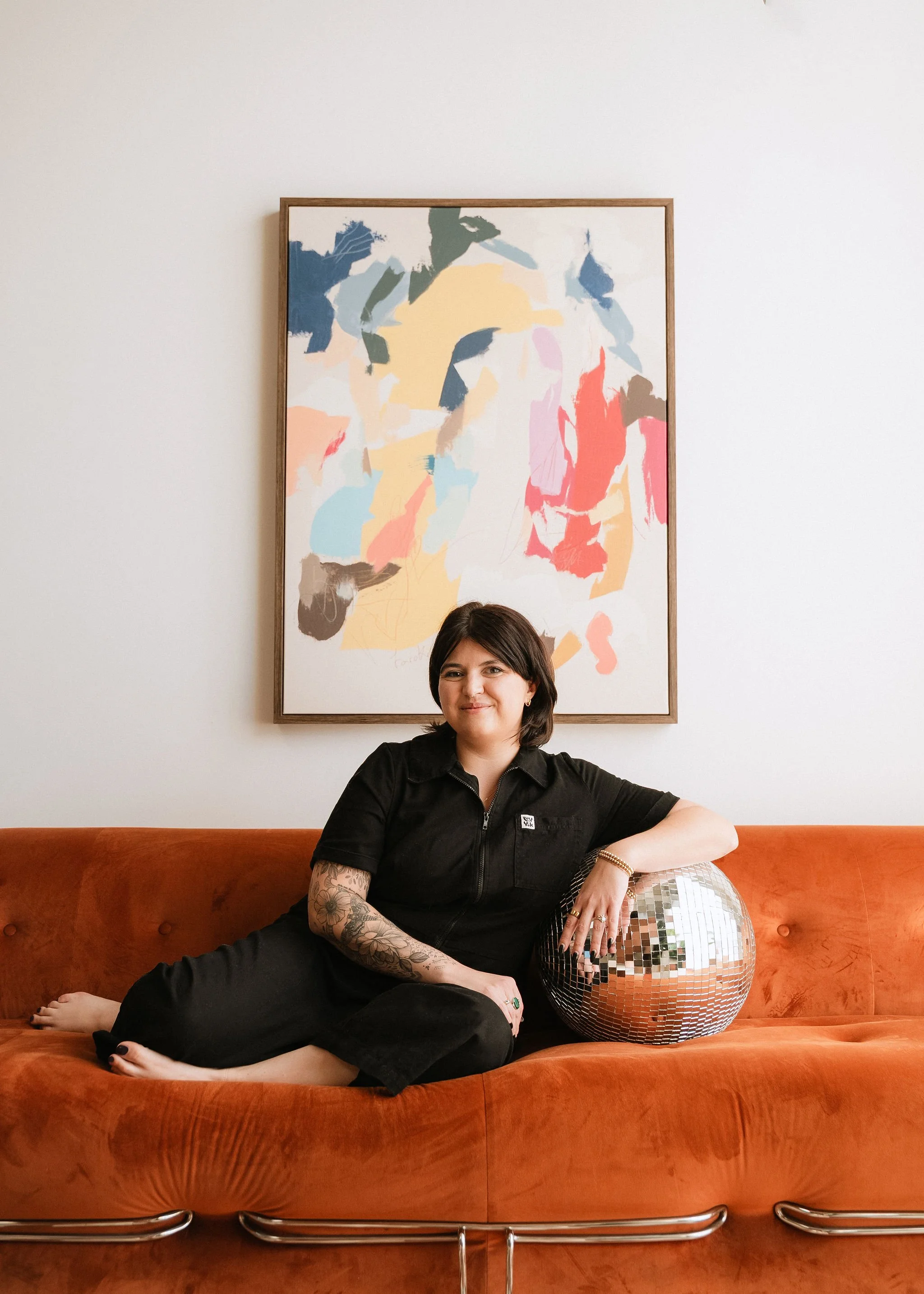 A woman with dark hair and tattoos, wearing a black Zipper hoodie, relaxing on an orange velvet couch with a disco ball, in front of a colorful abstract painting on a white wall.