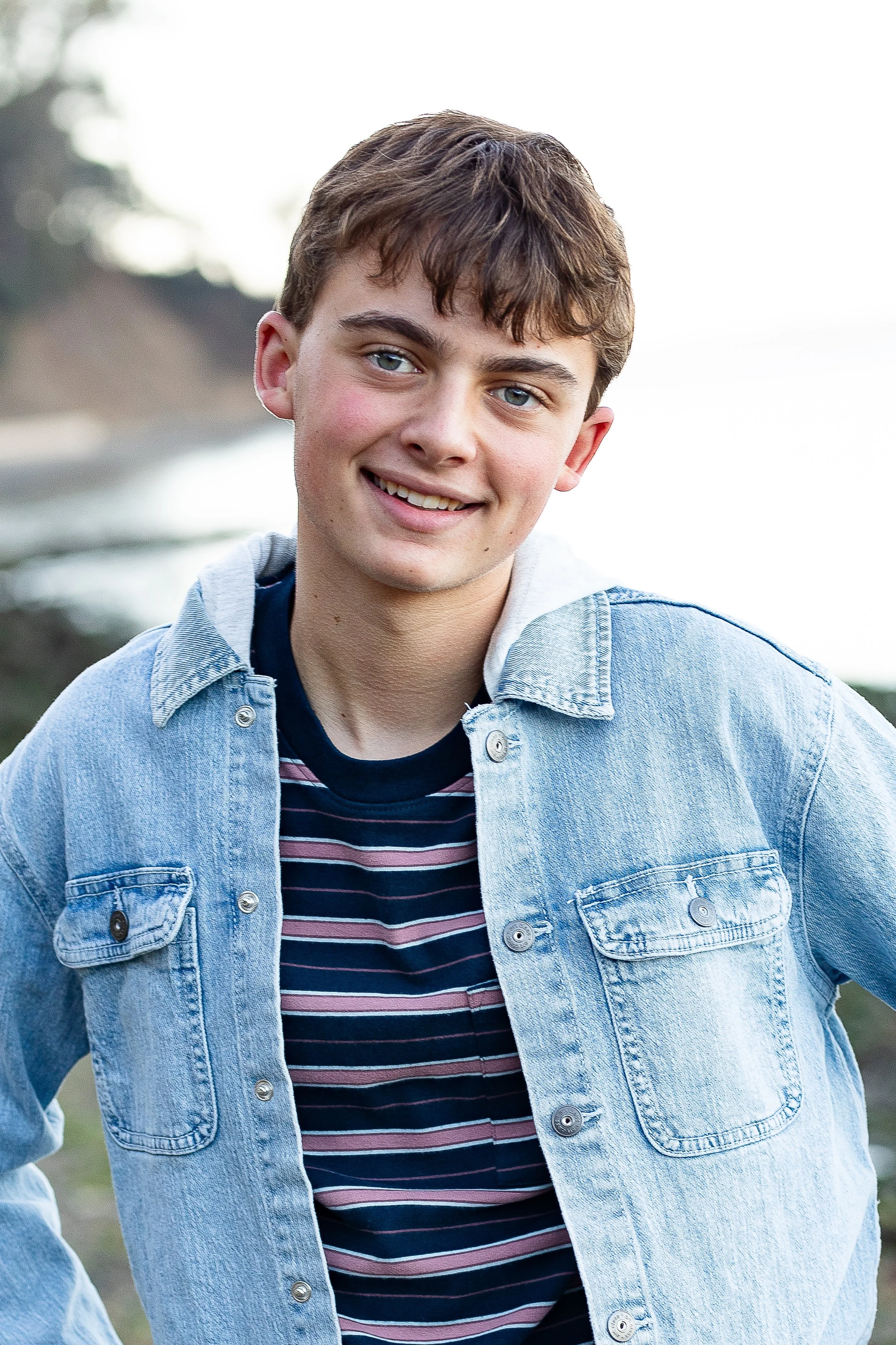 A young man smiling outdoors near water, wearing a striped t-shirt and denim jacket.