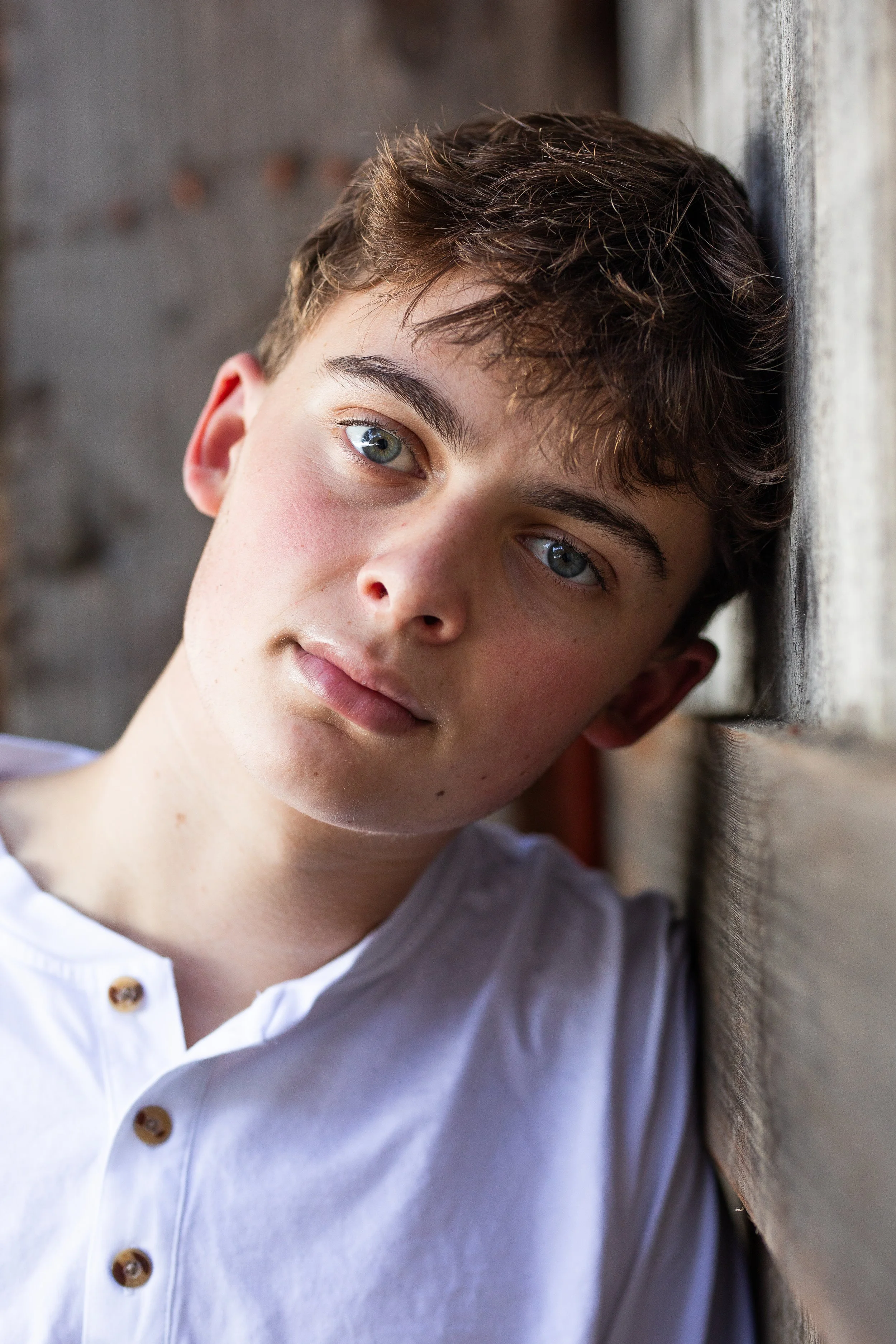 Close-up photo of a young man with light skin and blue eyes, leaning against a wooden wall, wearing a white shirt with brown buttons.