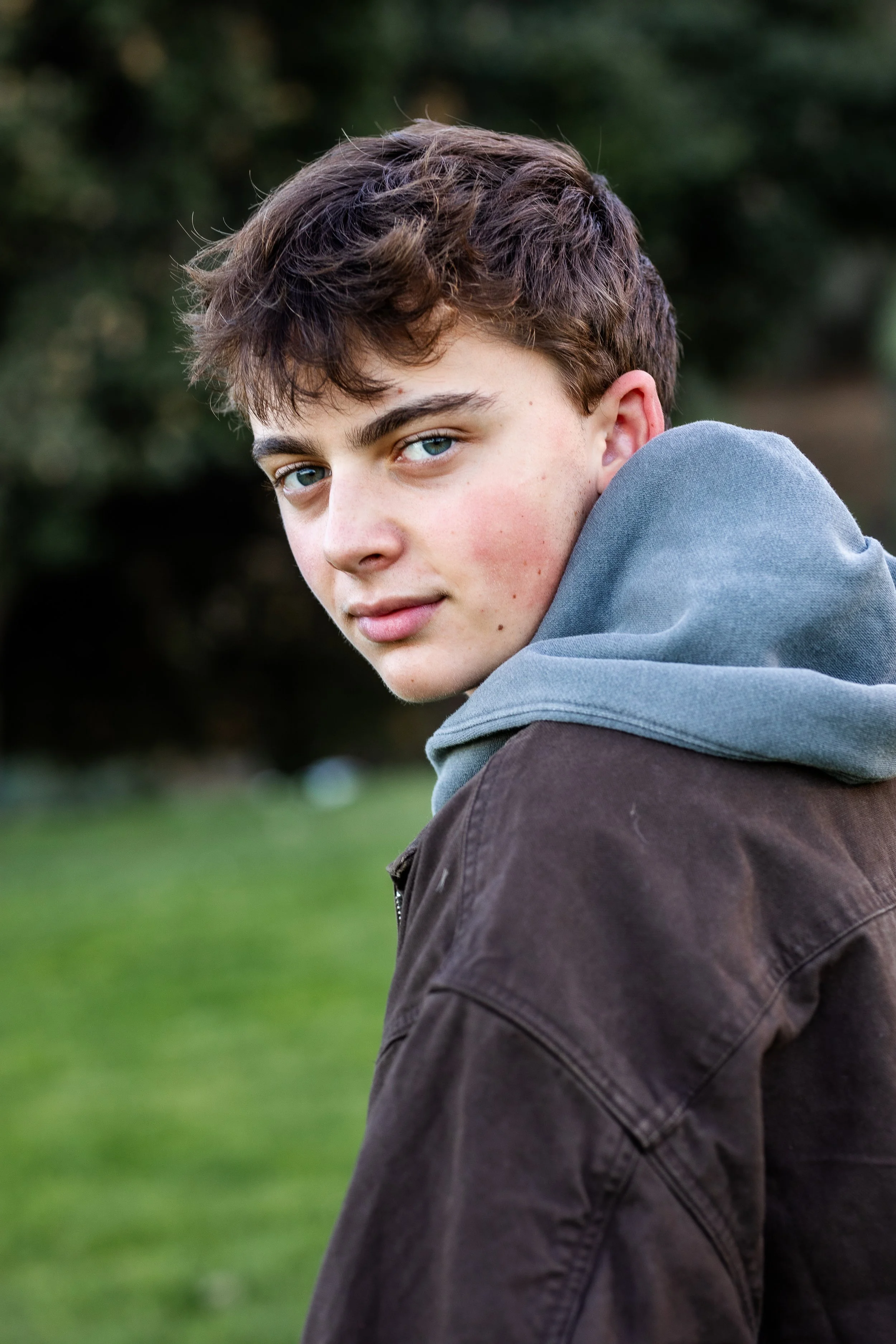 A teenage boy with brown hair and blue eyes, wearing a gray hoodie and brown jacket, looking back over his shoulder outdoors.