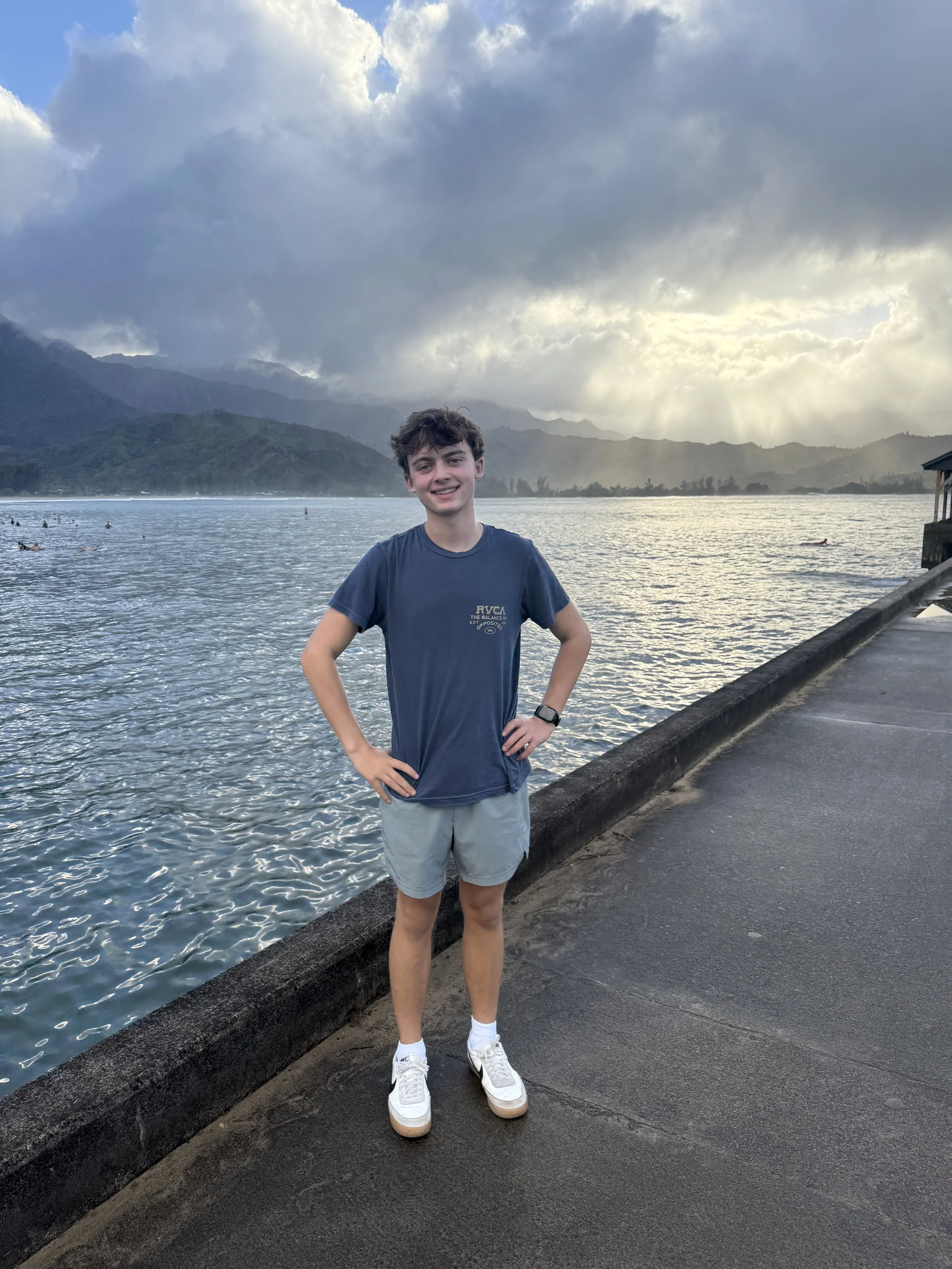 A young man standing on a waterfront promenade with mountains and a cloudy sky in the background at sunset.
