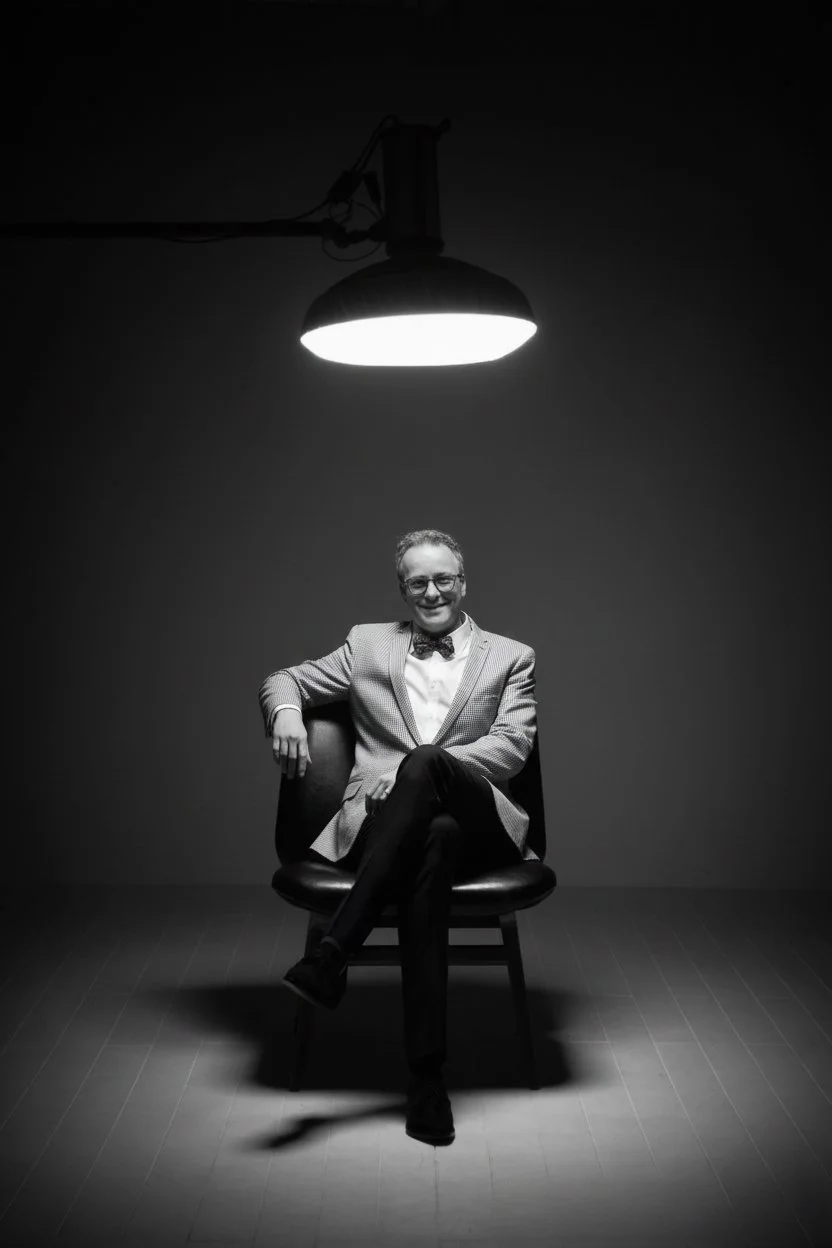 A man in a blazer and bowtie sitting on a chair in a dimly lit room with a large overhead light fixture.