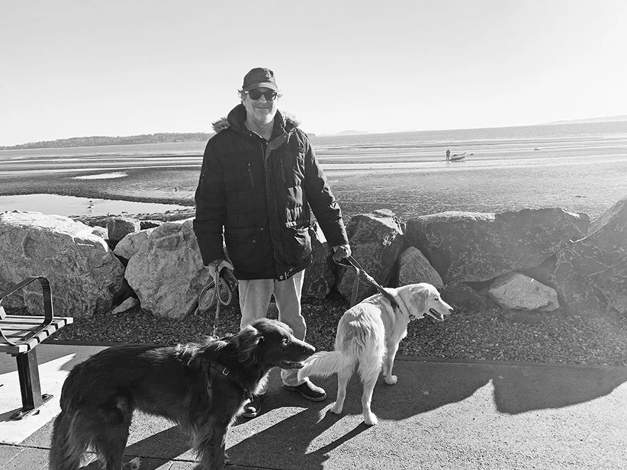 A person standing outdoors on a coastal walkway with two dogs. The person is wearing a hat, sunglasses, a jacket, and light-colored pants. One dog is dark-colored and the other is light-colored, both on leashes. The background features rocks, sand, water, and a distant shoreline.