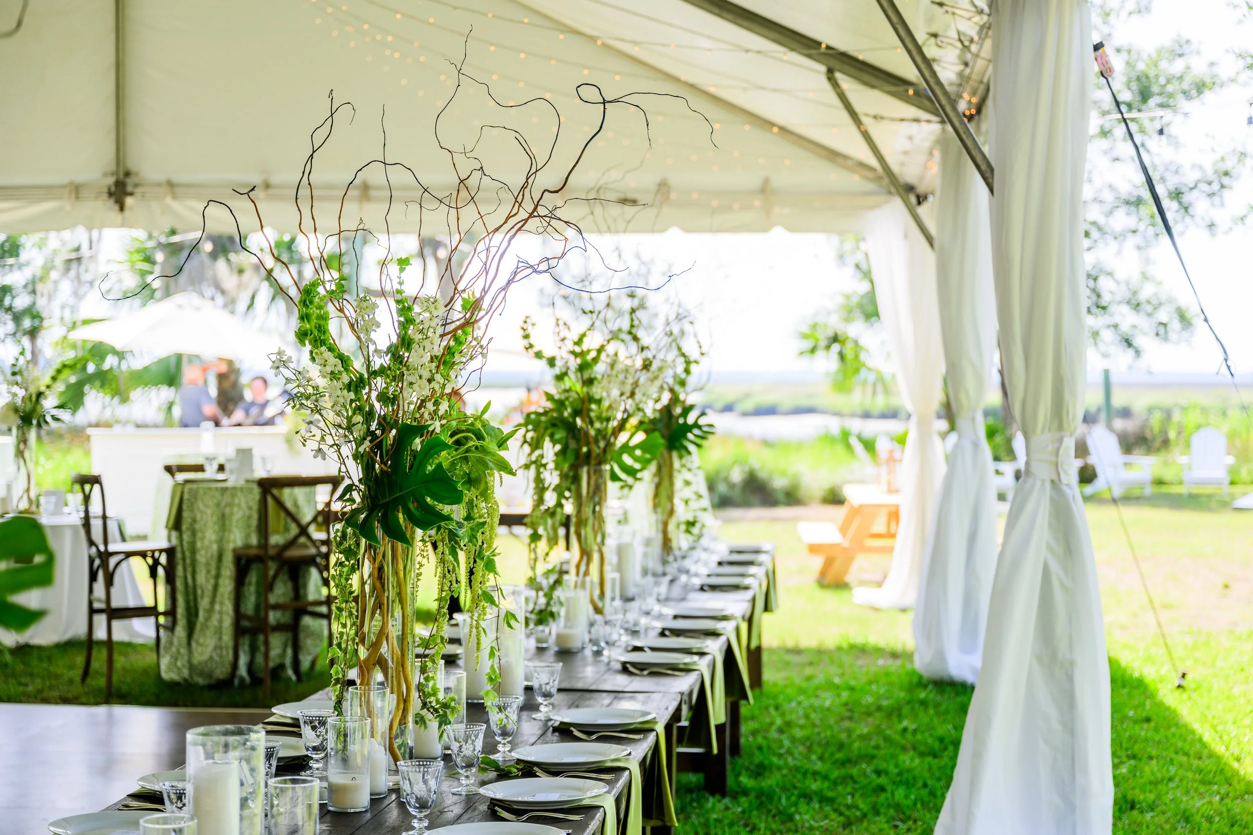 Decorated outdoor wedding or event setup under a white tent, featuring a long table with chairs, floral centerpieces, glassware, and candles, with string lights hanging from the ceiling and greenery visible in the background.