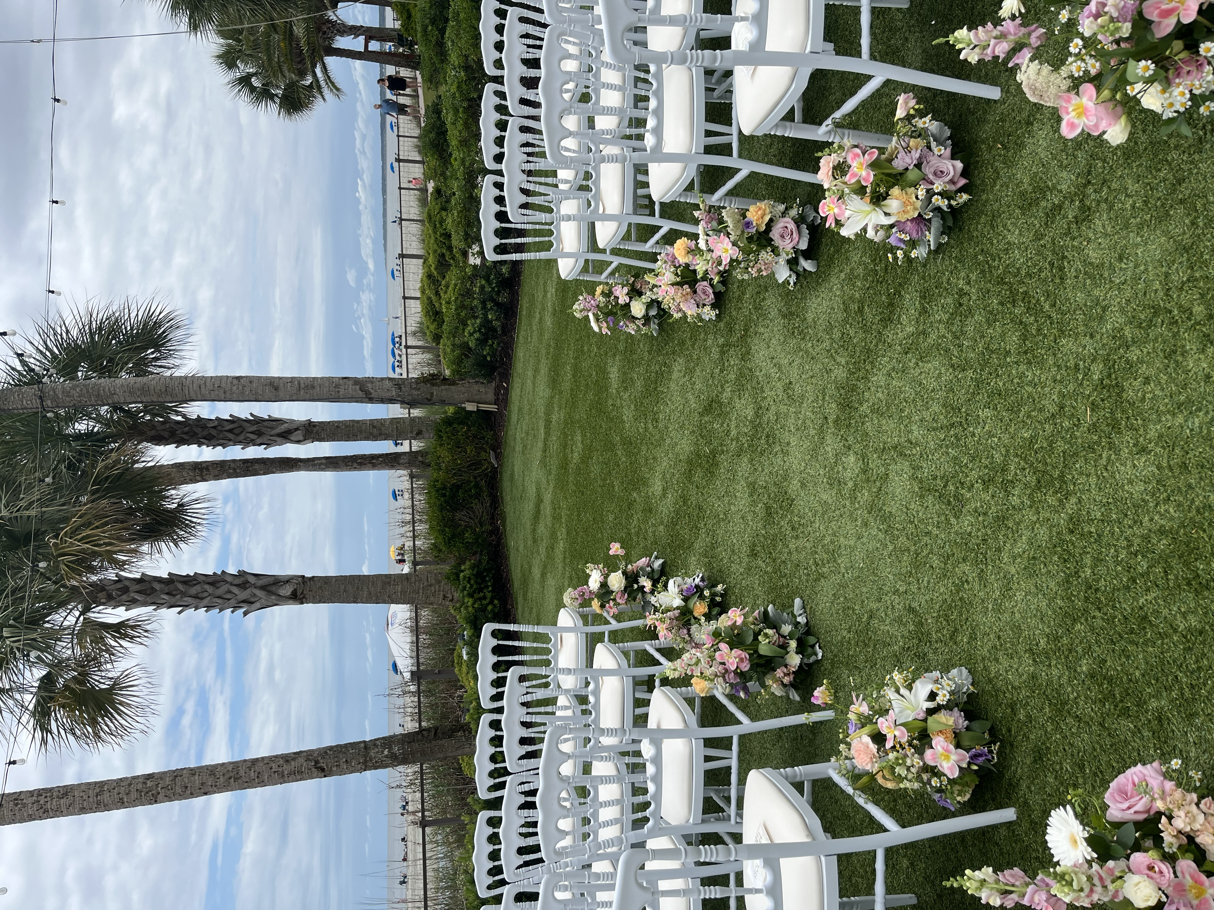 White chairs arranged outdoors on a grassy lawn, decorated with pink, white, and purple flowers, with palm trees and a cloudy sky in the background.