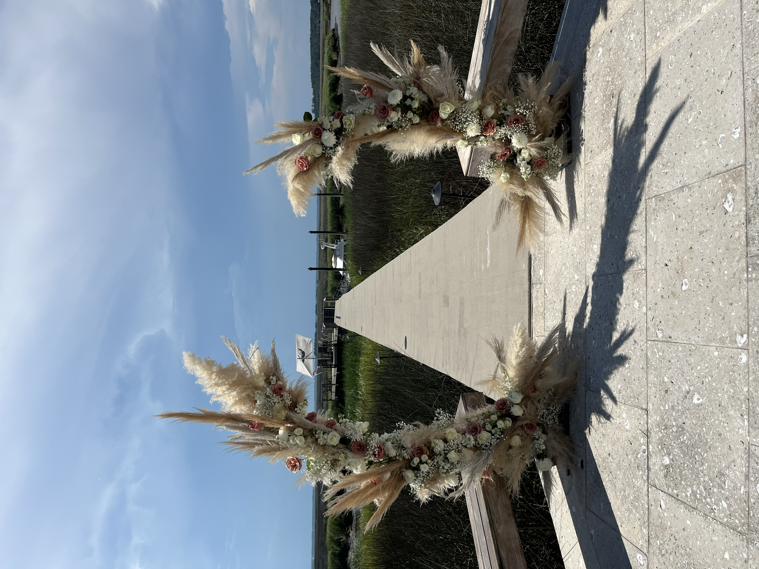 Outdoor floral arrangement with pampas grass and roses on a wooden structure by a lake with a cloudy sky.