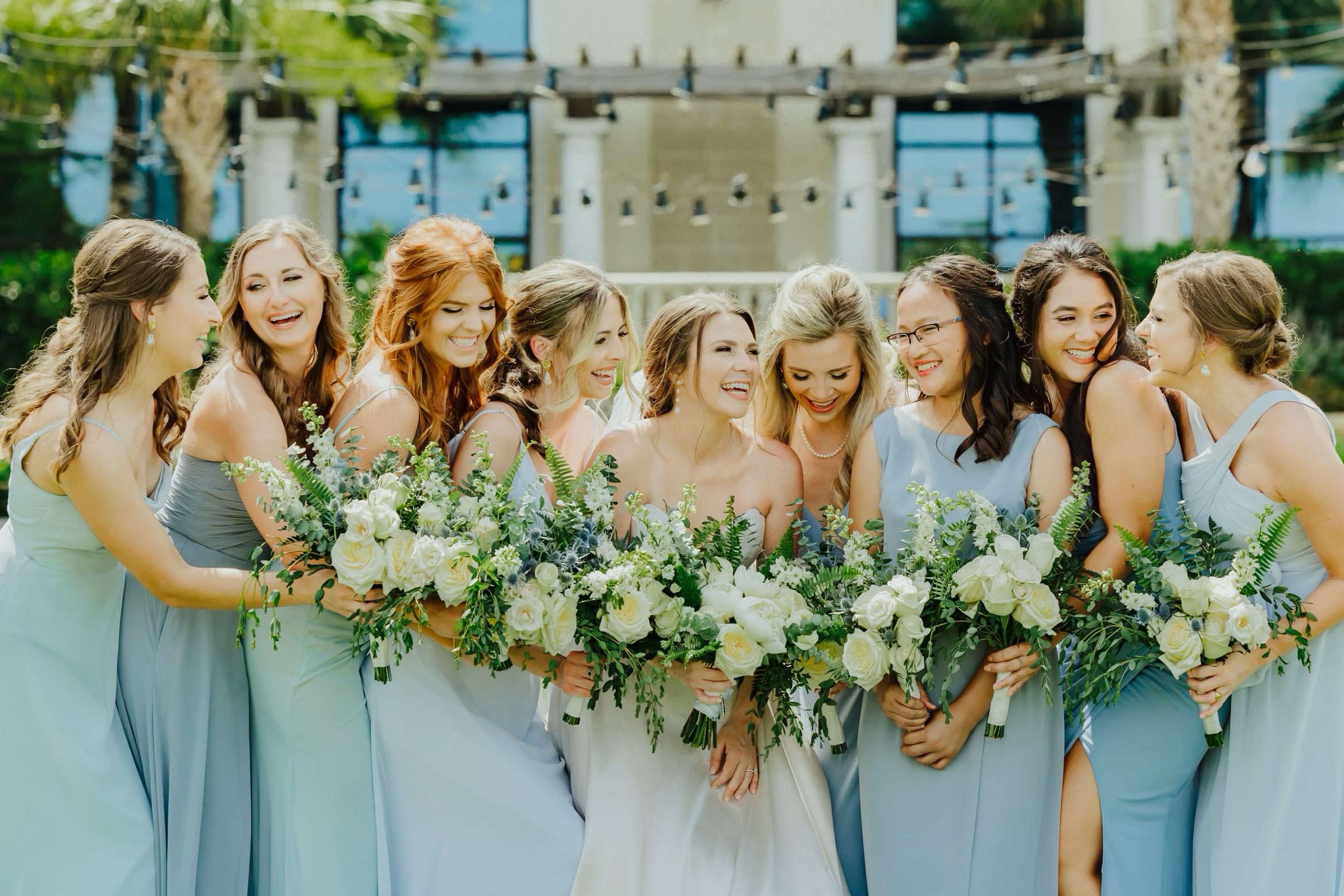 A group of women in bridesmaid dresses standing closely together outdoors, smiling and holding bouquets of white flowers and greenery, at a wedding or celebration.