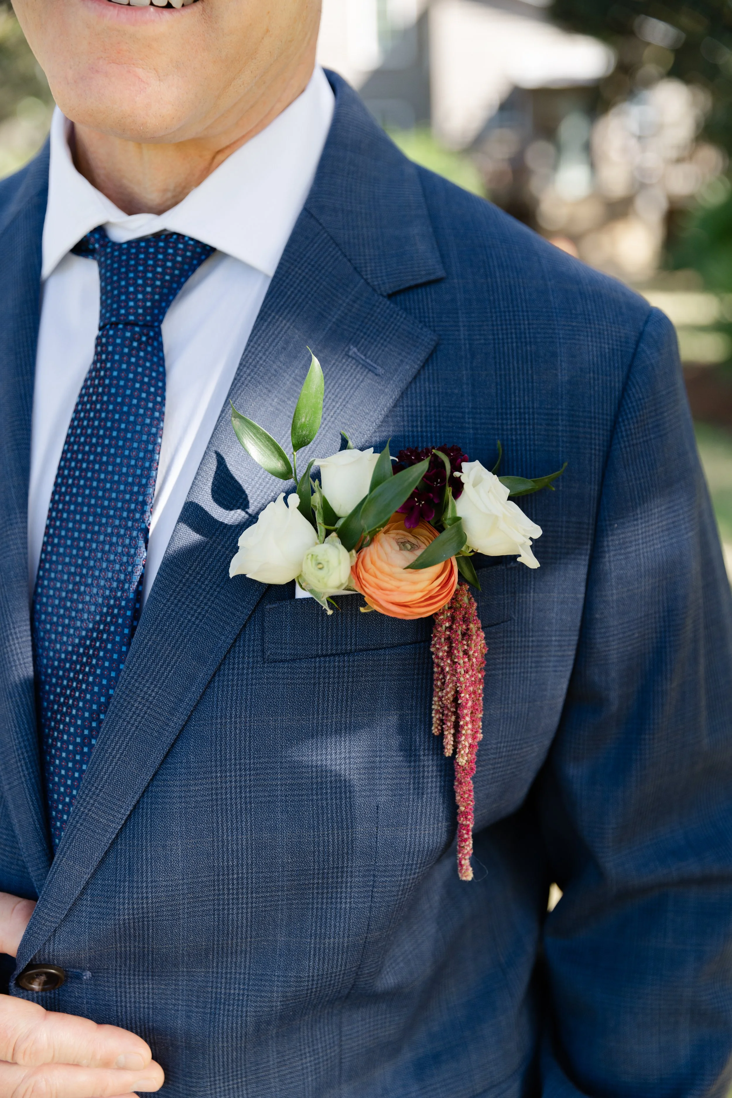 Close-up of a man in a blue suit jacket, white shirt, and patterned blue tie, with a boutonniere featuring white, orange, and dark purple flowers, and pink hanging amaranthus on his lapel. He is smiling slightly.