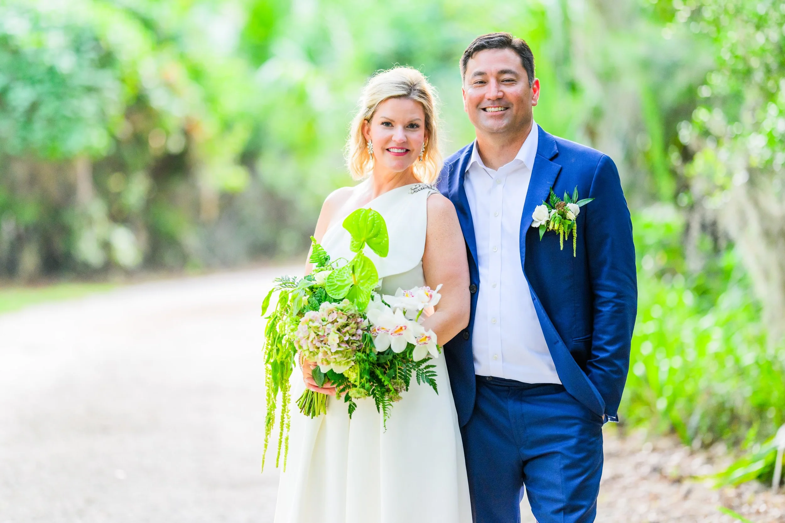 A smiling couple, a woman wearing a white dress and a man in a blue suit, standing outdoors in a lush green park during daytime. The woman is holding a bouquet of flowers.