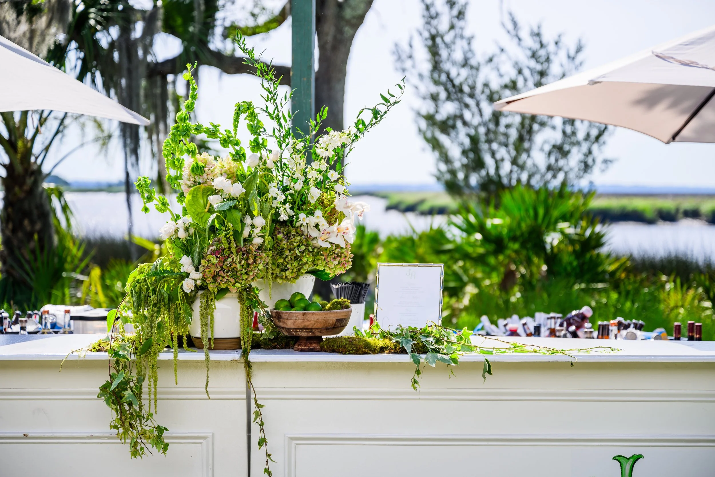 A white table with a tall floral arrangement of white and green flowers, surrounded by bottles, and set outdoors with umbrellas and trees in the background.