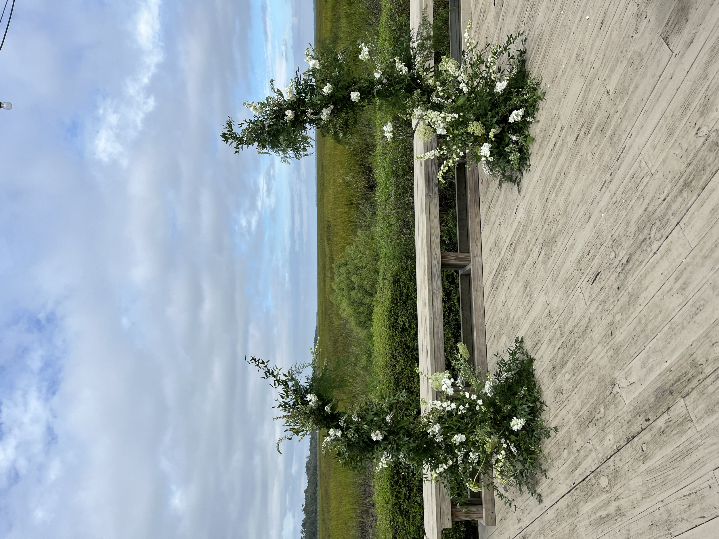 A large flower arrangement with white flowers and green foliage on a wooden deck outdoors. In the background, there is a wide field with grass and shrubs, and a cloudy sky overhead.