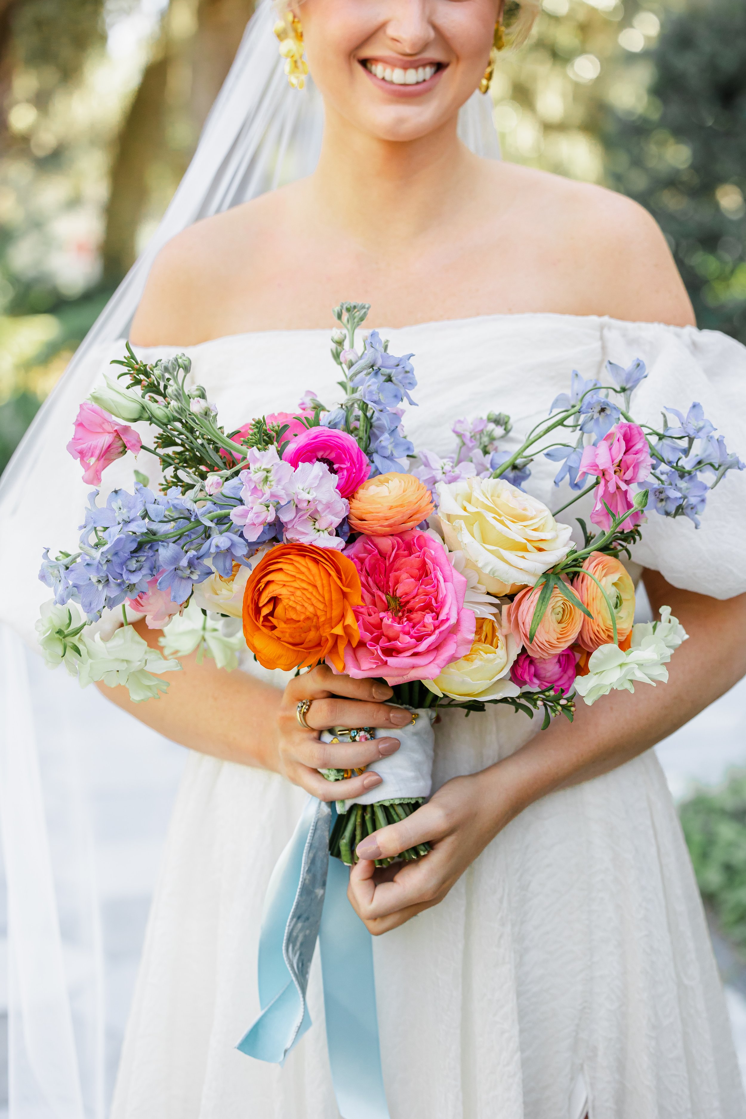 Close-up of a smiling woman in a white off-shoulder dress holding a colorful bouquet of flowers, with a blurred outdoor background.