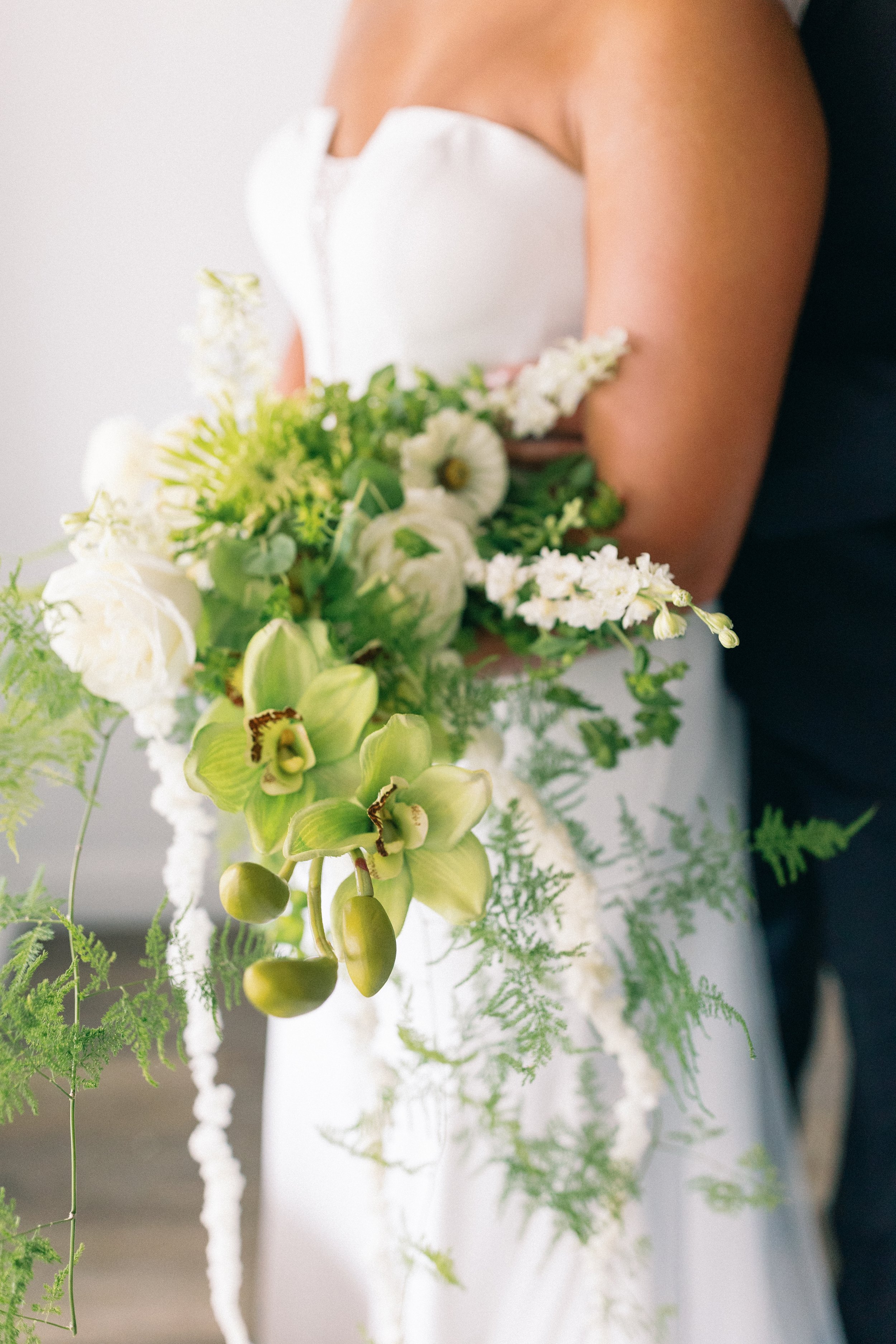Close-up of a woman holding a bouquet of white and green flowers, wearing a strapless white dress, with a blurred background.