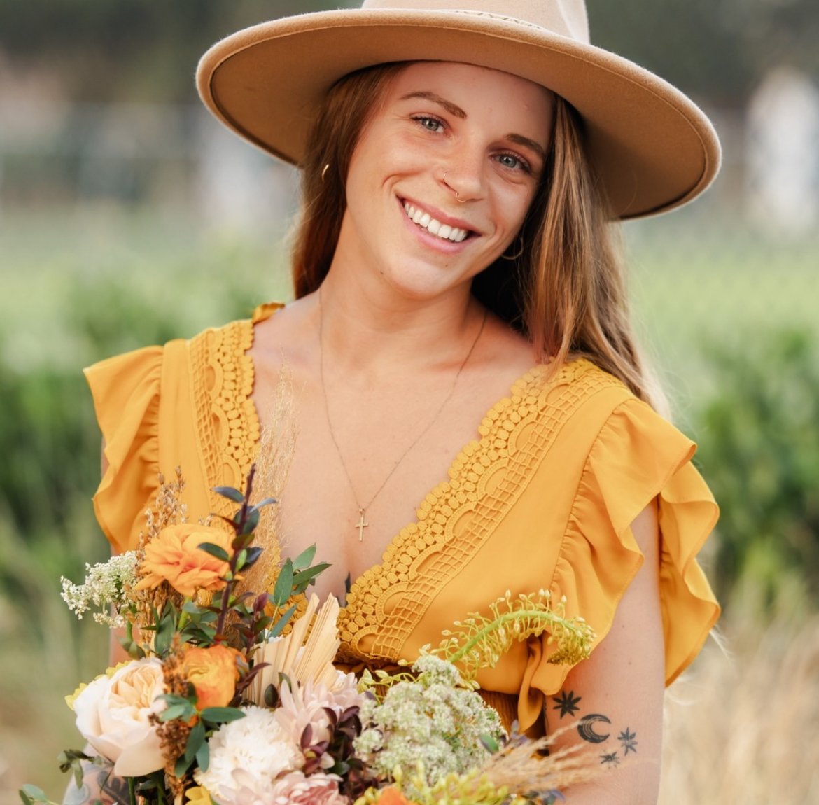 A woman smiling outdoors, wearing a wide-brimmed hat and a yellow dress with lace details, holding a bouquet of flowers.