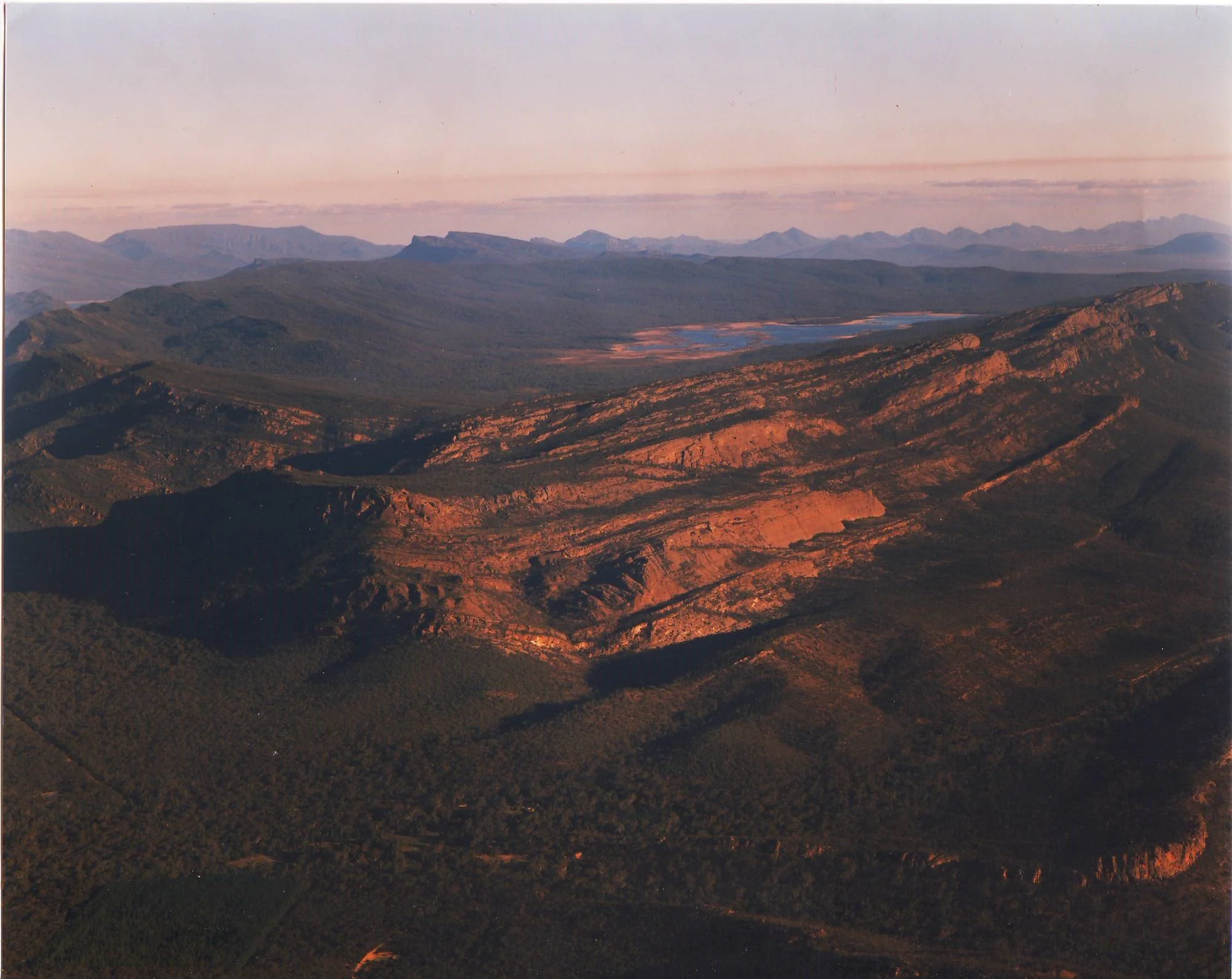 A vast mountain range at sunset with rocky peaks, green valleys, and a small lake in the distance.