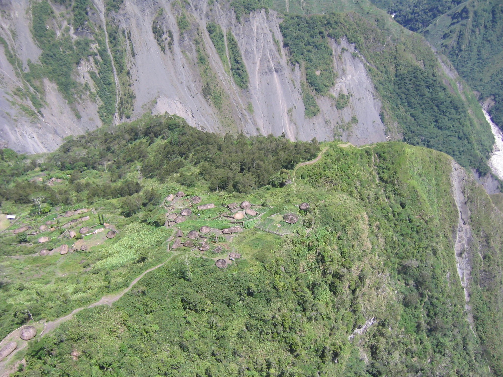 A green mountainside with a small village of thatched-roof huts and a winding dirt path.