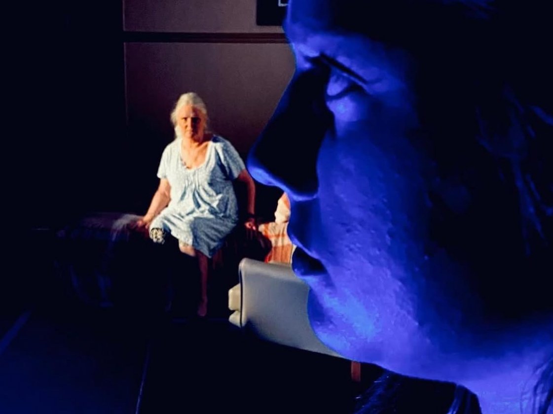 A woman in a hospital gown sitting on a chair, with a person in blue lighting in close profile foreground.