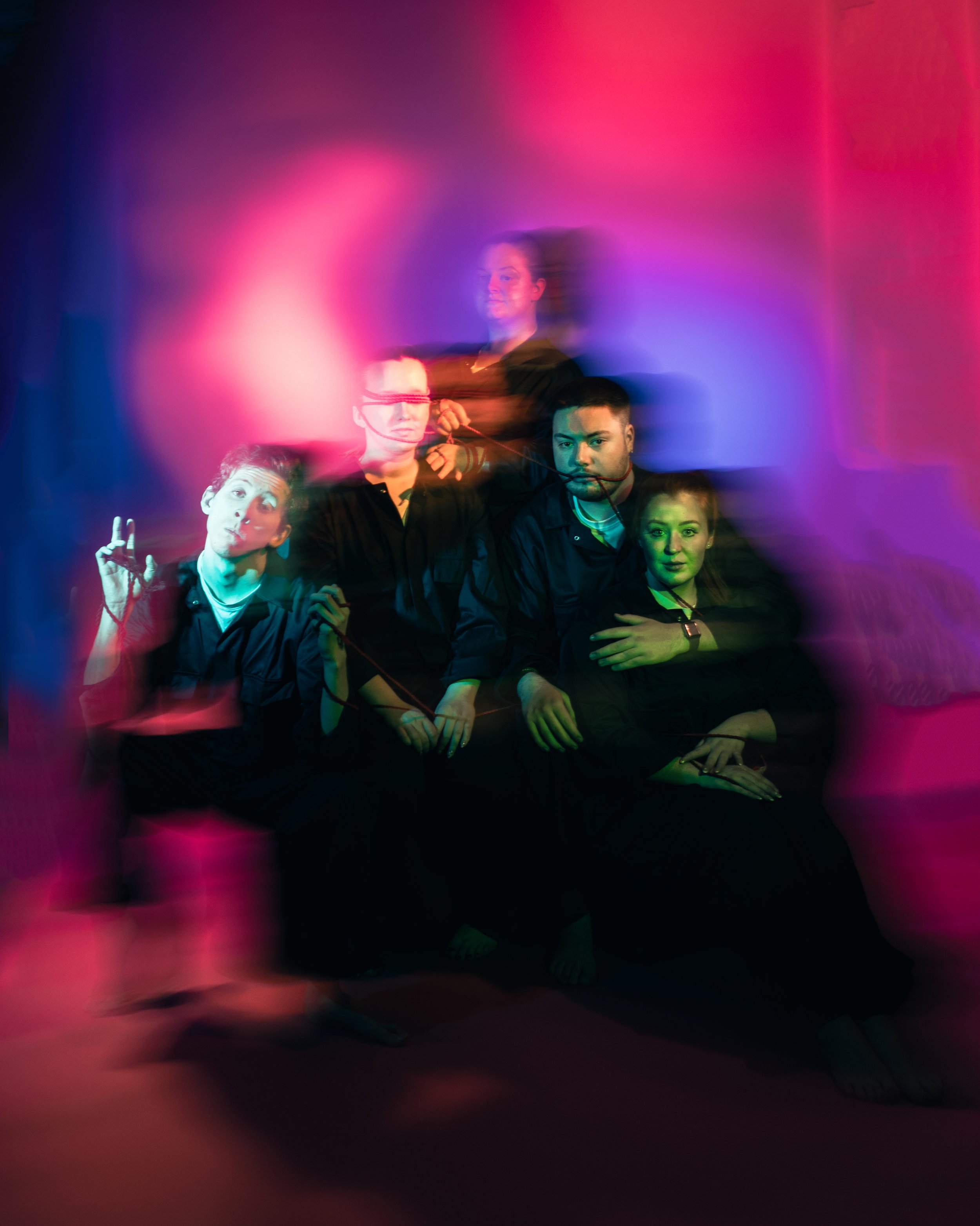 Group of five young adults sitting on a red floor in a dark room with colorful, blurred light trails creating a vibrant, abstract background.