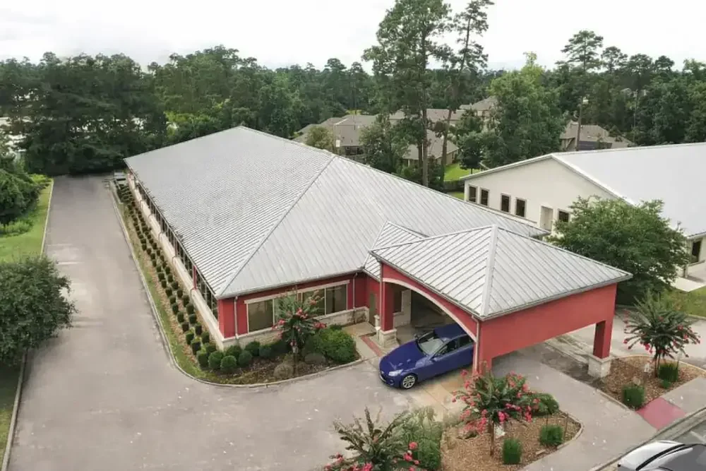 An aerial view of a single-story building with a metal roof, red walls, and a covered driveway with a blue car parked underneath. The building is surrounded by landscaped bushes and trees.