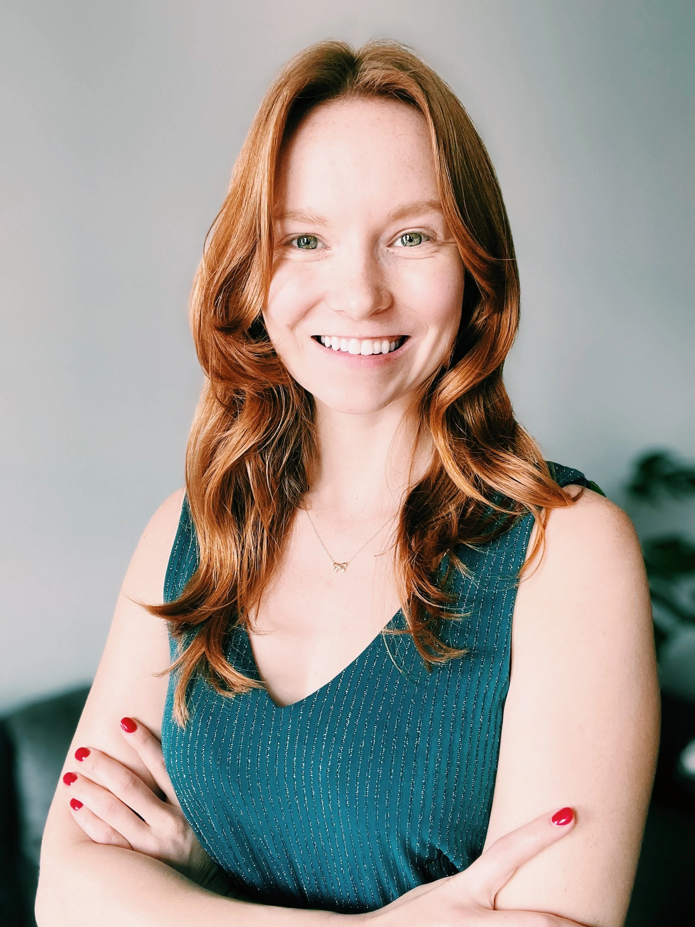 A smiling woman with red hair, crossed arms, wearing a sleeveless teal top and a delicate necklace, standing indoors with a light-colored wall and a blurred plant in the background.