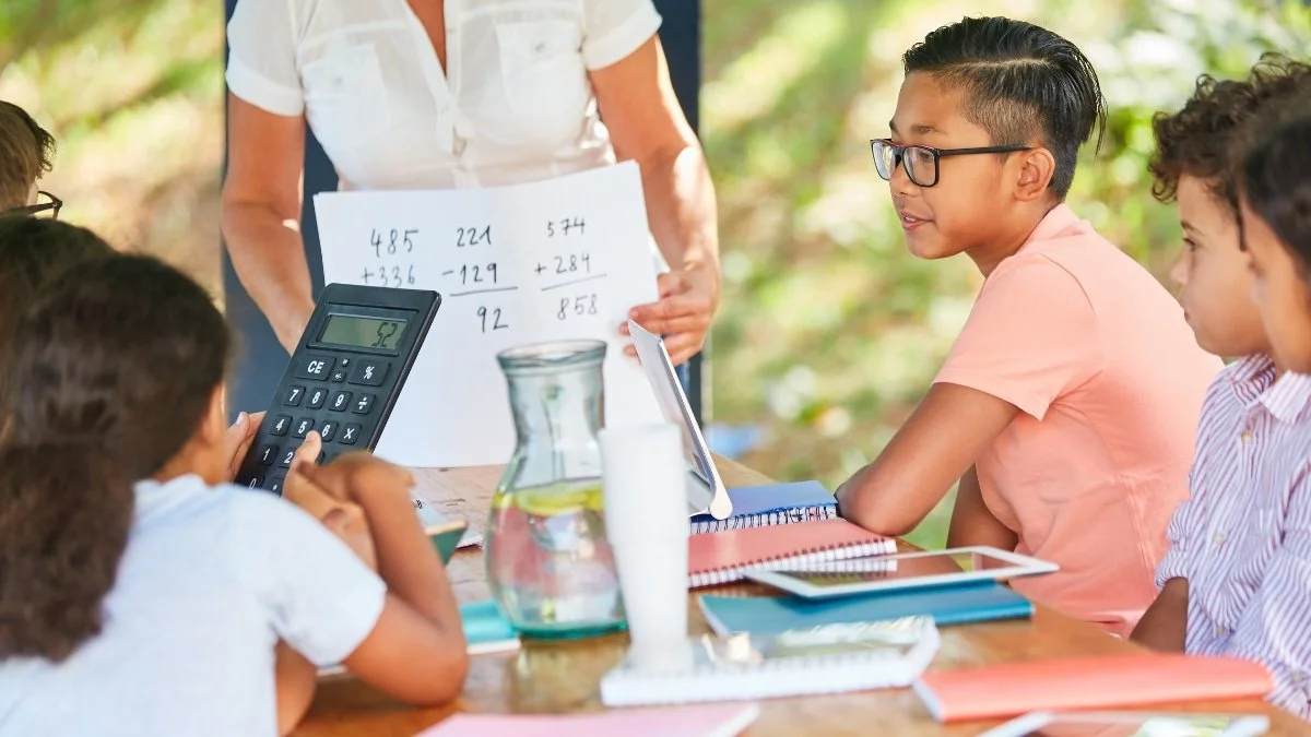 Six kids at a table with their teacher learning math for a blogpost about economics for kids.