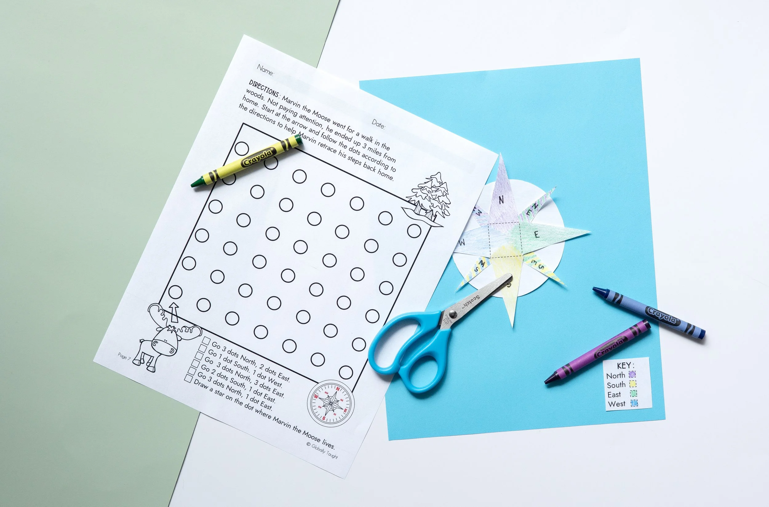 A compass rose worksheet and craft from Globally Taught's World Geography Unit, with a Crayola-green crayon, blue scissors, a purple Crayola crayon, and a paper compass rose with labeled directions and a key for directions.
