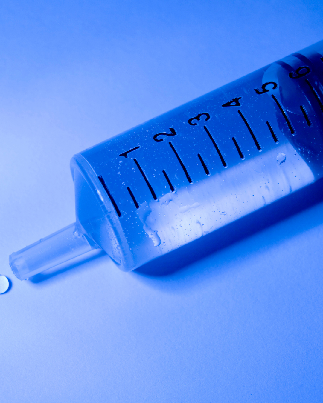 A close-up of a medical syringe laying diagonally on a light blue surface, with visible measurement markings and droplets of liquid on the transparent plastic.